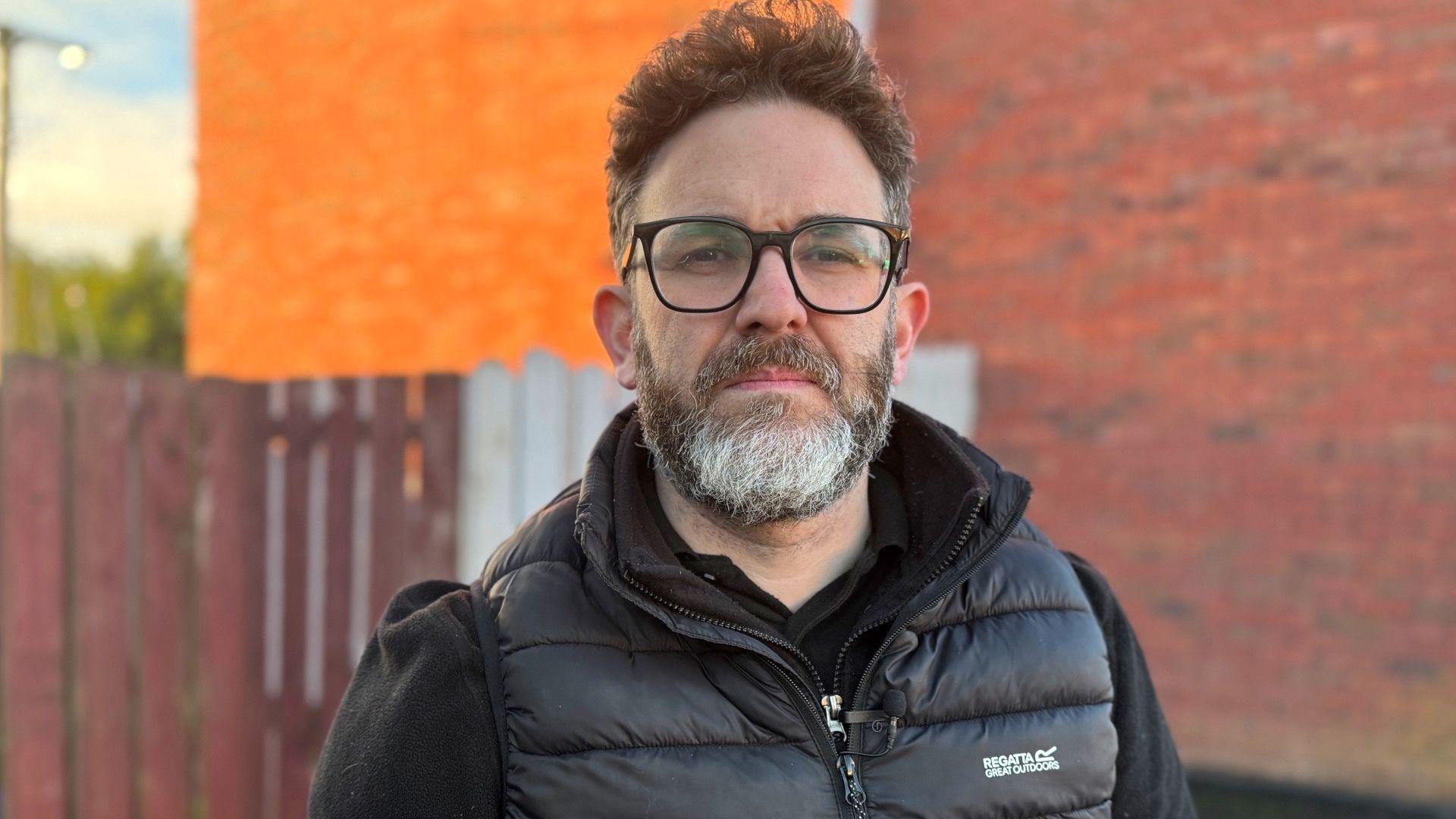 A man with dark hair and a dark grey beard looks toward the camera. He is wearing a black waterproof jacket and black rimmed glasses. In the background is an exterior wall of a red brick house and a brown fence.