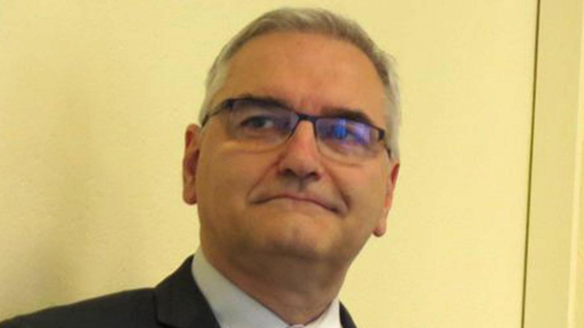 A man in a dark suit jacket, pale shirt and blue tie with short grey hair. He is wearing glasses. There is a plain cream-coloured wall behind him.