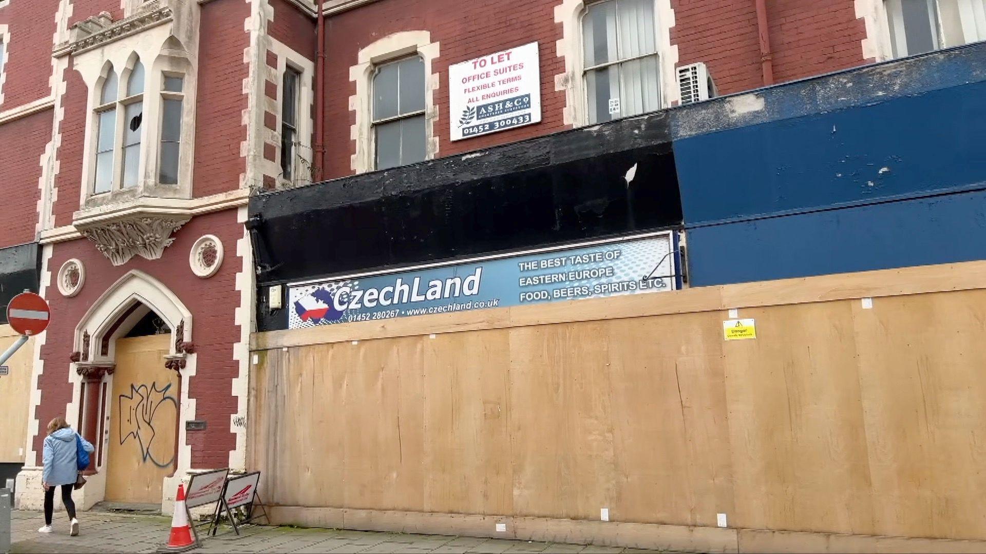 A boarded up building with an ornate red brick frontage with white features is the main subject of this photo. The the building looks dilapidated and one of the first floor windows has been broken. There's graffiti on one of the boarded up panels and a person walking past the front of he building. There's a 'to let' sign on the right hand side of the building.