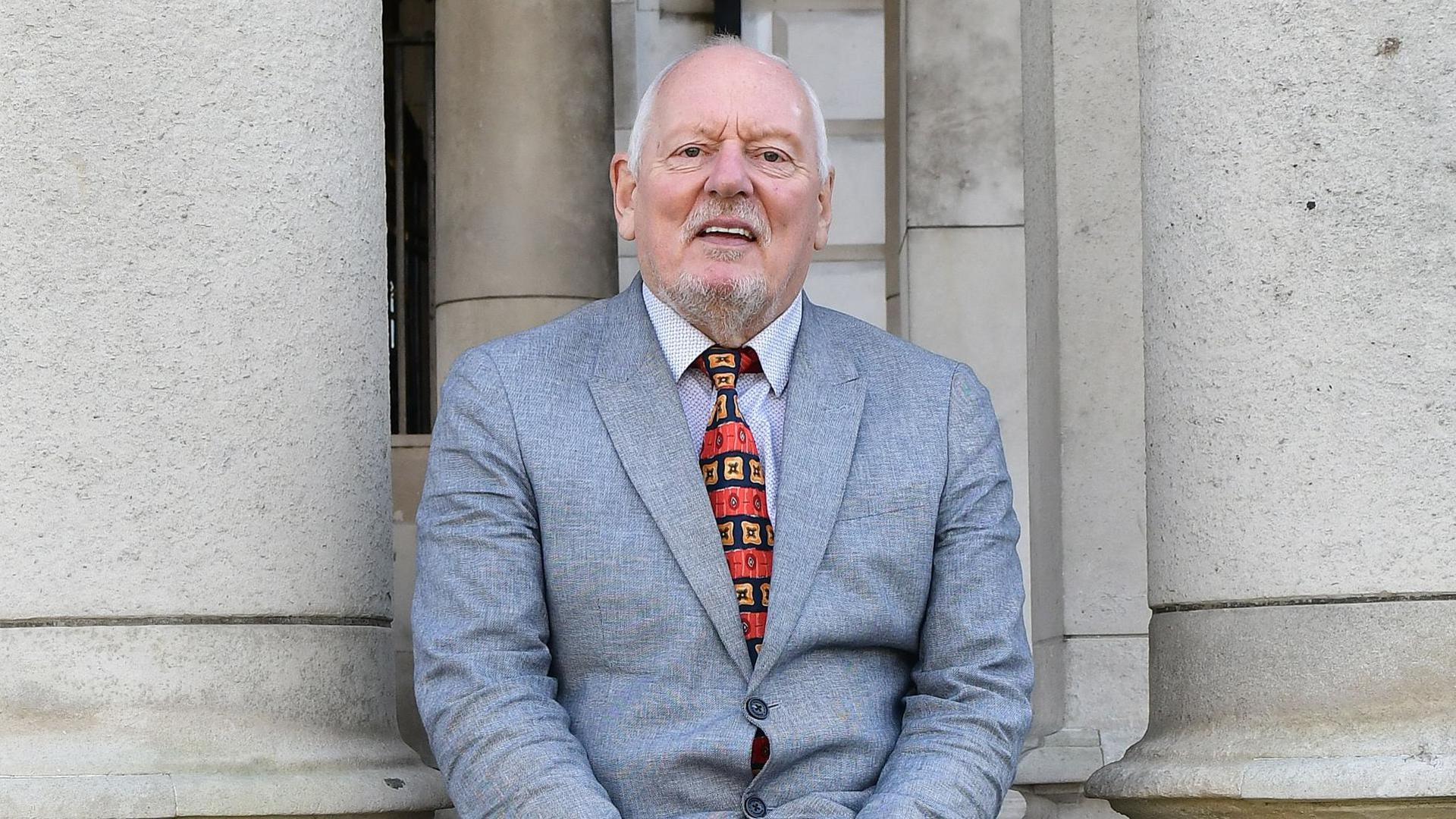 Jeff Dudgeon sitting outside Belfast City Hall in 2019.  He has white hair and a goatee beard.  He is wearing a light grey suit jacket and a red and gold patterned tie. 