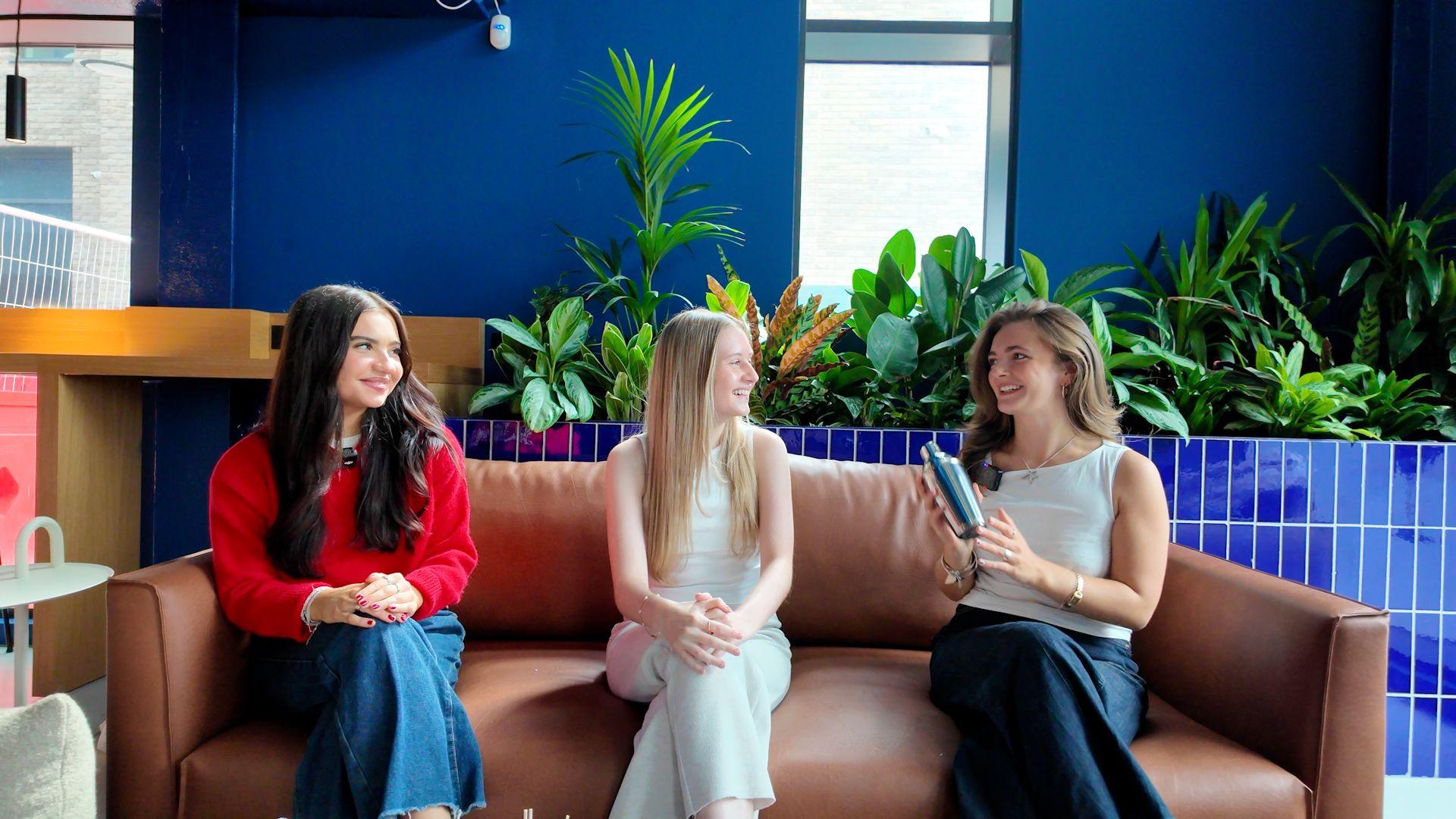 Three students sat on a brown sofa. The student on the right is showing the other two a cocktail shaker