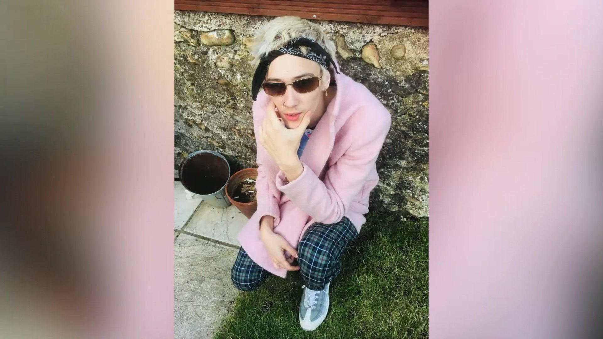 A young boy with a pink blazer, chequered trousers, and shades squats striking a pose in a garden. 