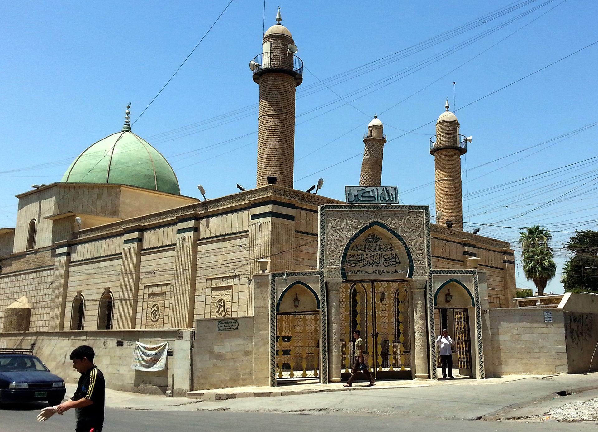 People walk in front of the Al-Noori Al-Kabeer mosque in Mosul. The green dome of the mosque is visible behind the main gate, with the leaning minaret poking above the building in the background against a blue, cloudless sky.