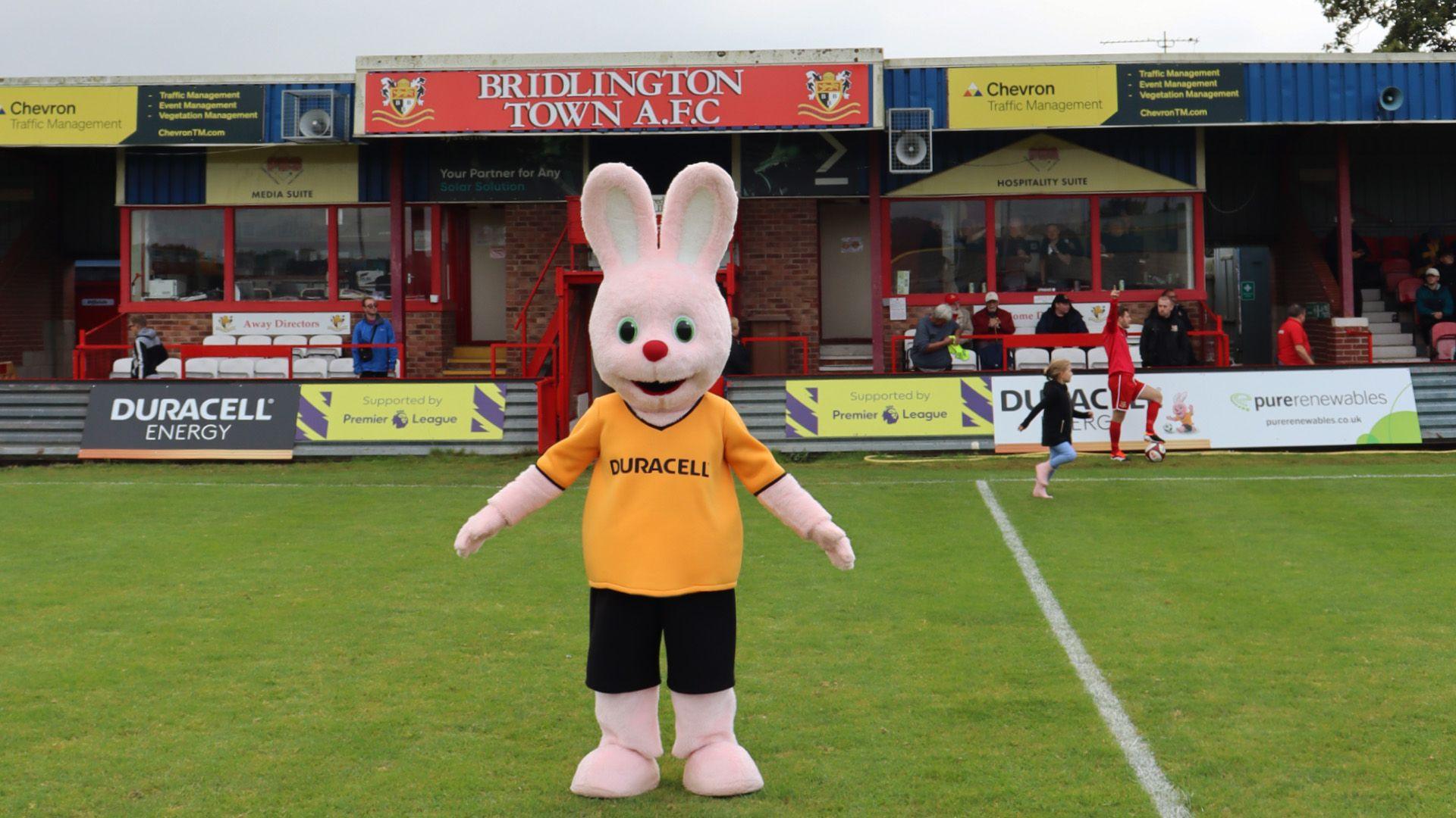 A person wearing a yellow and black Duracell bunny outfit standing on a green football pitch in front of a stand with a banner that reads: "Bridlington Town AFC".