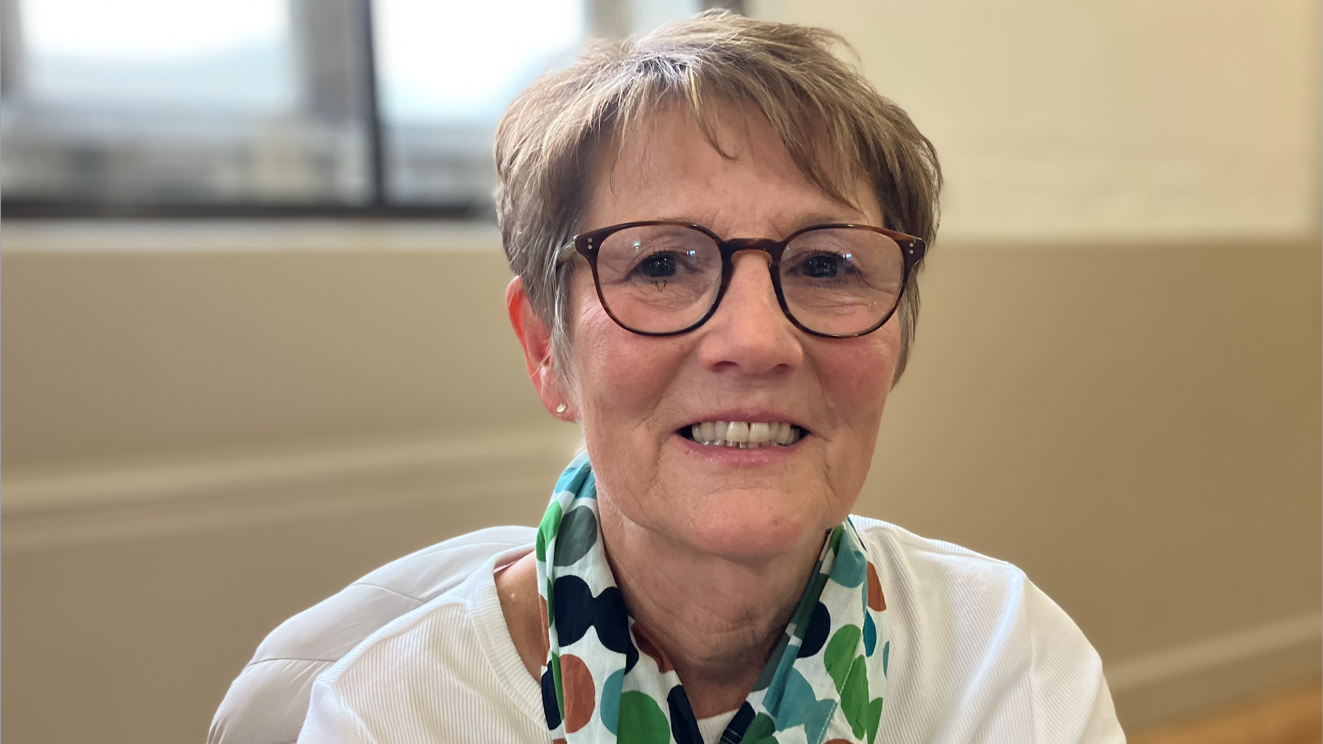 Angela Ellis, sitting down, looking at the camera and smiling, wearing glasses with short fair hair. She has a scarf round her neck and a white top on. She is in a room with cream walls behind her. 