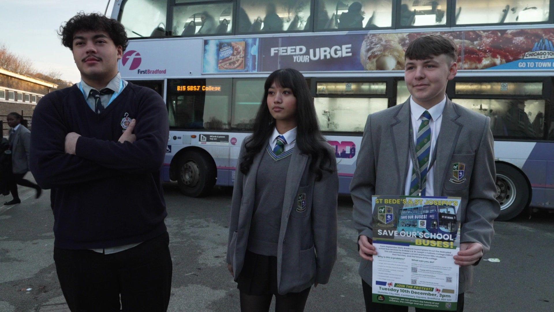 Three teenagers in school uniform, two male and one female. One of the male students is holding a poster which says 'Save Our School Buses'. A school bus is parked behind them.