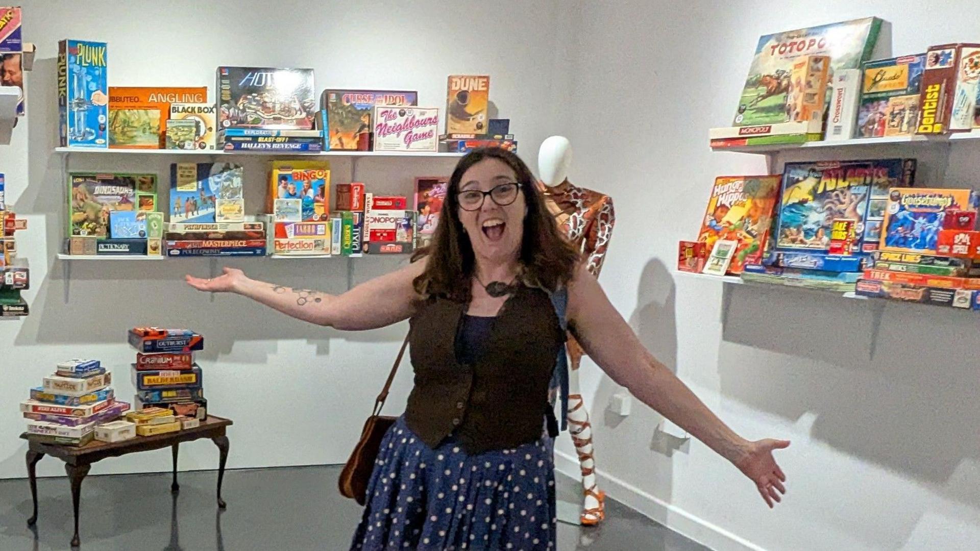 Ann, with long black hair and glasses. She is smiling and gesturing her arms to the area packed with about 60 games behind her.