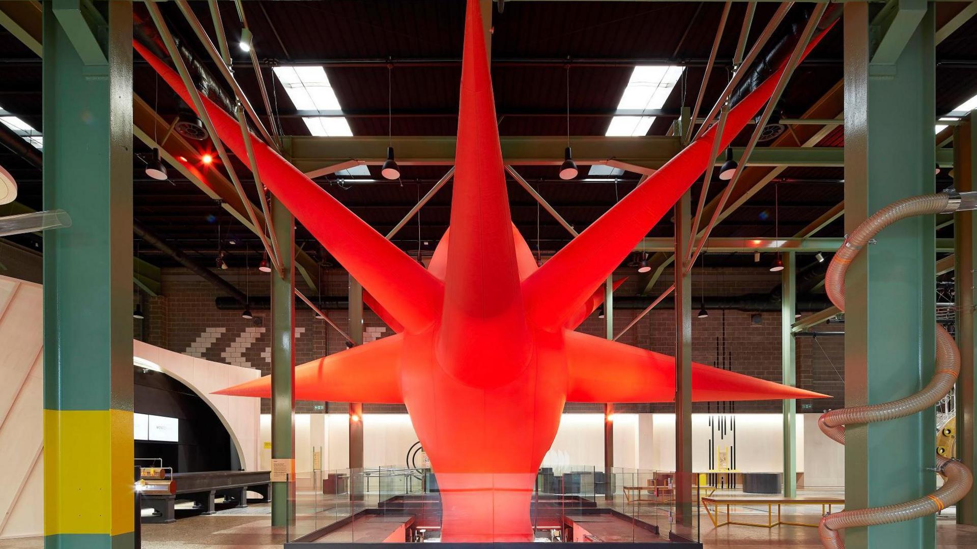 A big, red oval-shaped structure with eight spikes emerging at various points. It is situated in a big hall.