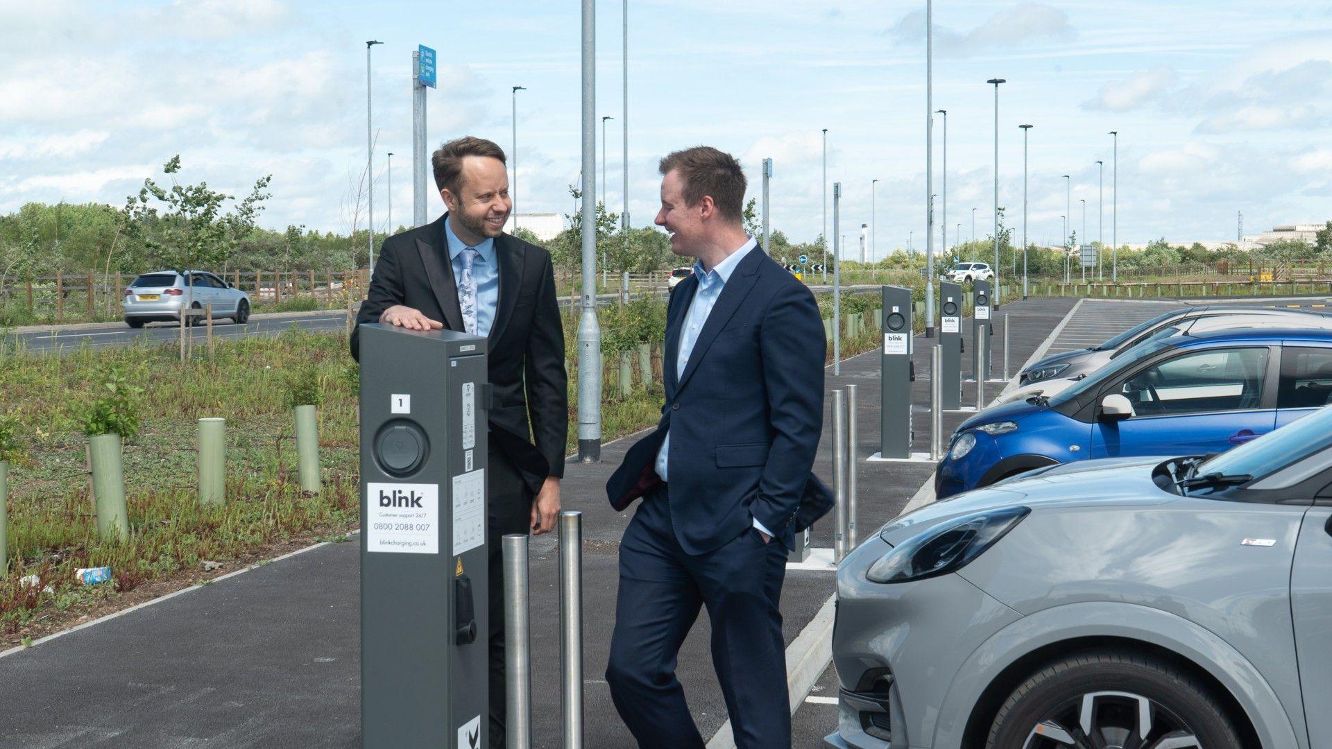 Two men in dark suits stand next to a grey car parked in a marked bay next to an EV charging post