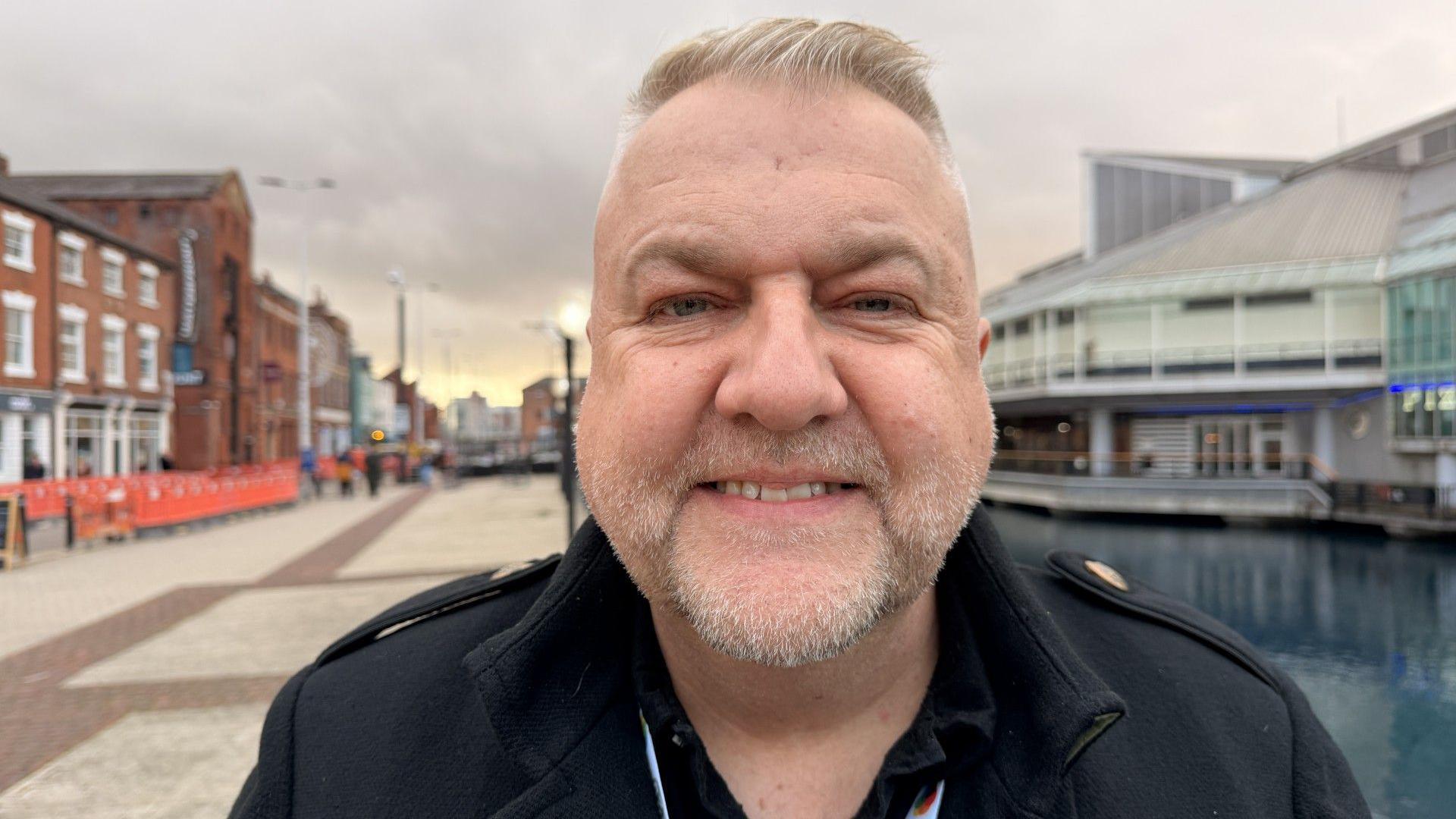 A head and shoulders image of a man standing on a dockside, with a wide promenade and converted warehouse buildings to the left and a dock and large 1980s shopping centre to the right. He has short blonde hair, swept back, stubble and is smiling. He is wearing a smart black coat.