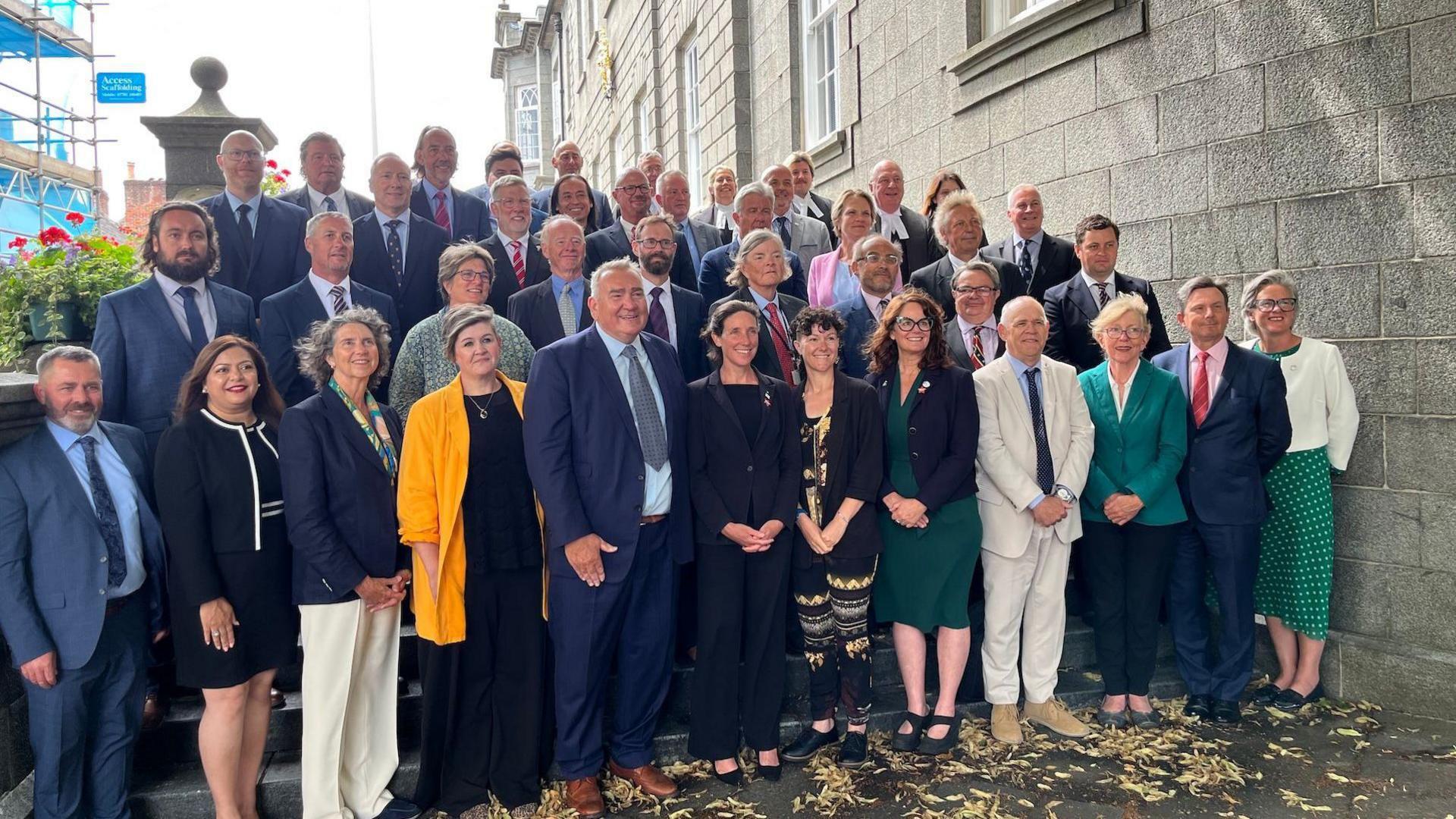 A large group of people stood on steps outside a large building. They are all dressed in smart suits, blazers and dresses and are all smiling looking towards a camera.