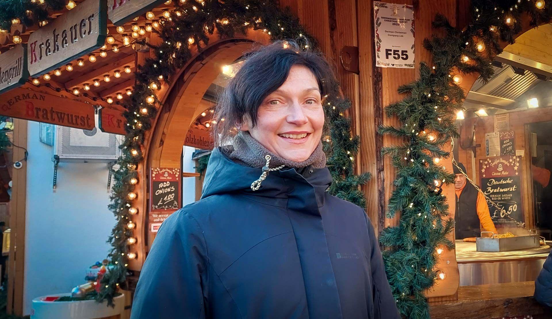 A woman with dark hair in a black coat stands in front of a wooden hut at a Christmas market, adorned with fake Christmas tree sprigs, fairylights, signs saying "Krakauer" and "Bratwurst"