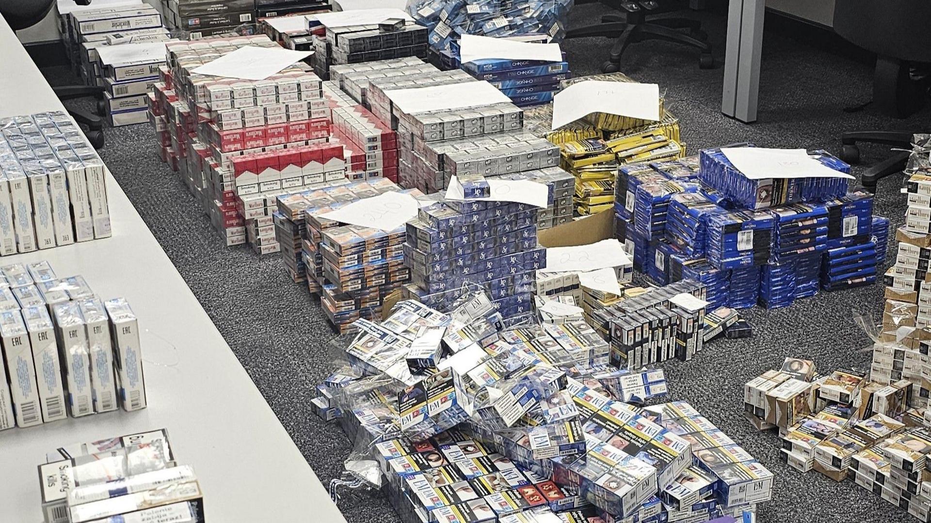 Hundreds of boxes of cigarettes and tobacco on the floor of an office. Some of the piles have sheets of paperwork on top of them.
