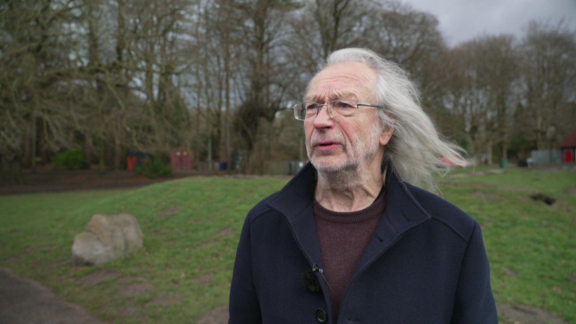 An elderly man with white long hair, blowing in the wind, and a white short beard, wearing glasses and a navy coat, frowns as he talks off to the left of the camera, standing in a park