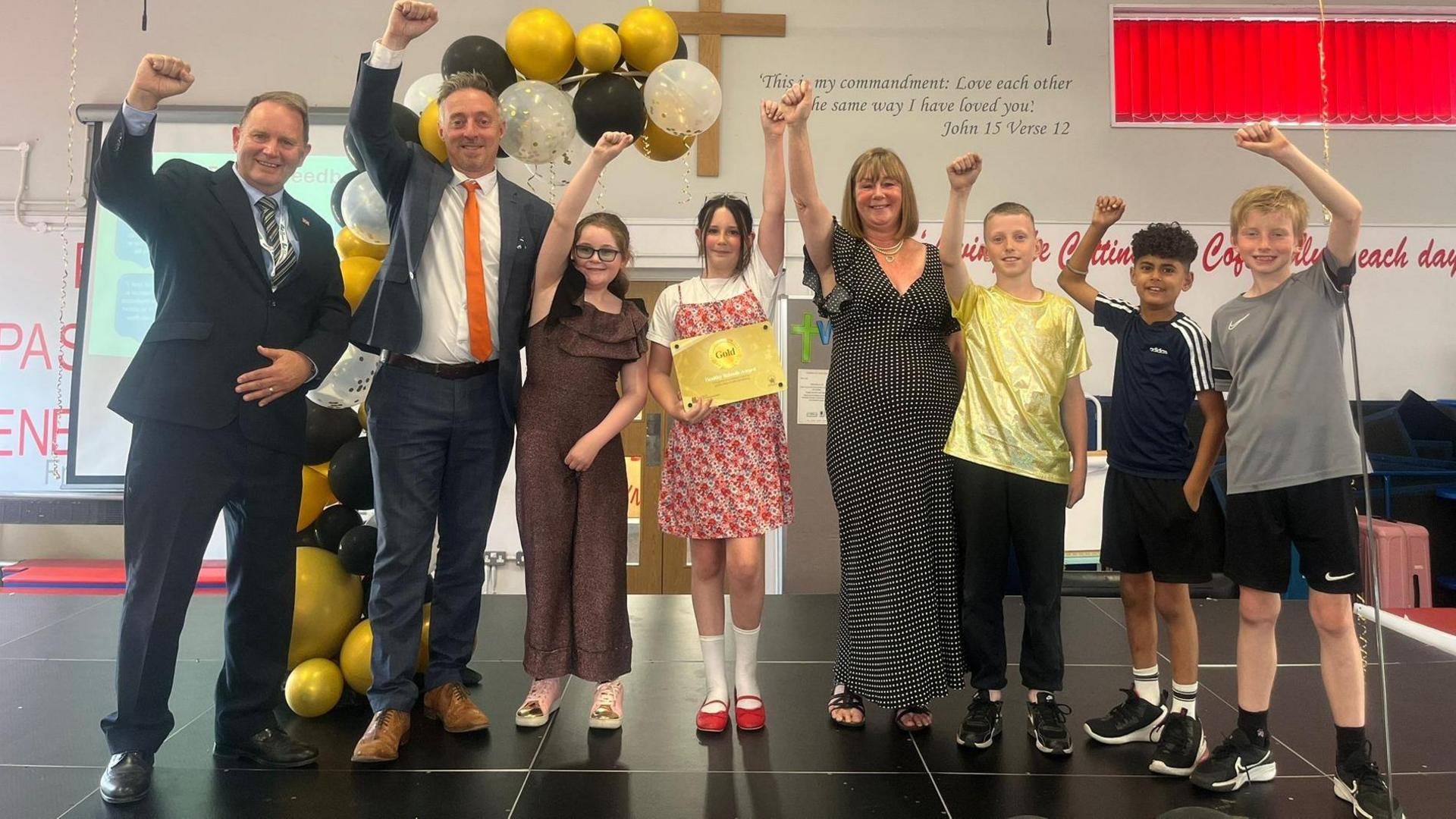 Eight people, all with their arms raised, looking happy and at the camera. Two men on the left are dressed in dark suits, with ties on, then five children are dressed in sports kits, one girl in a dress and one girl in a gold jumpsuit. A woman is wearing a black and white dress. 