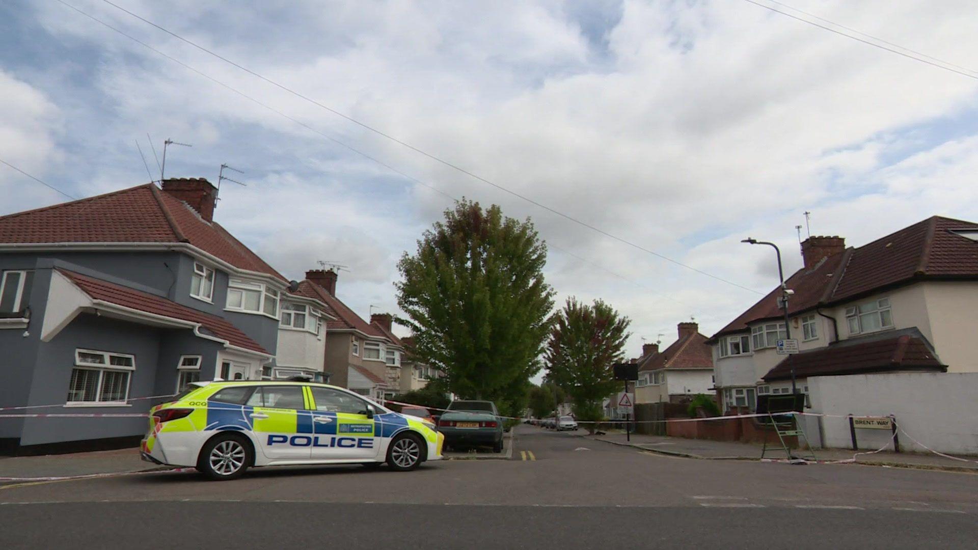 A police car parked at the end of a road, with a police tape cordon stretching across the road entrance.