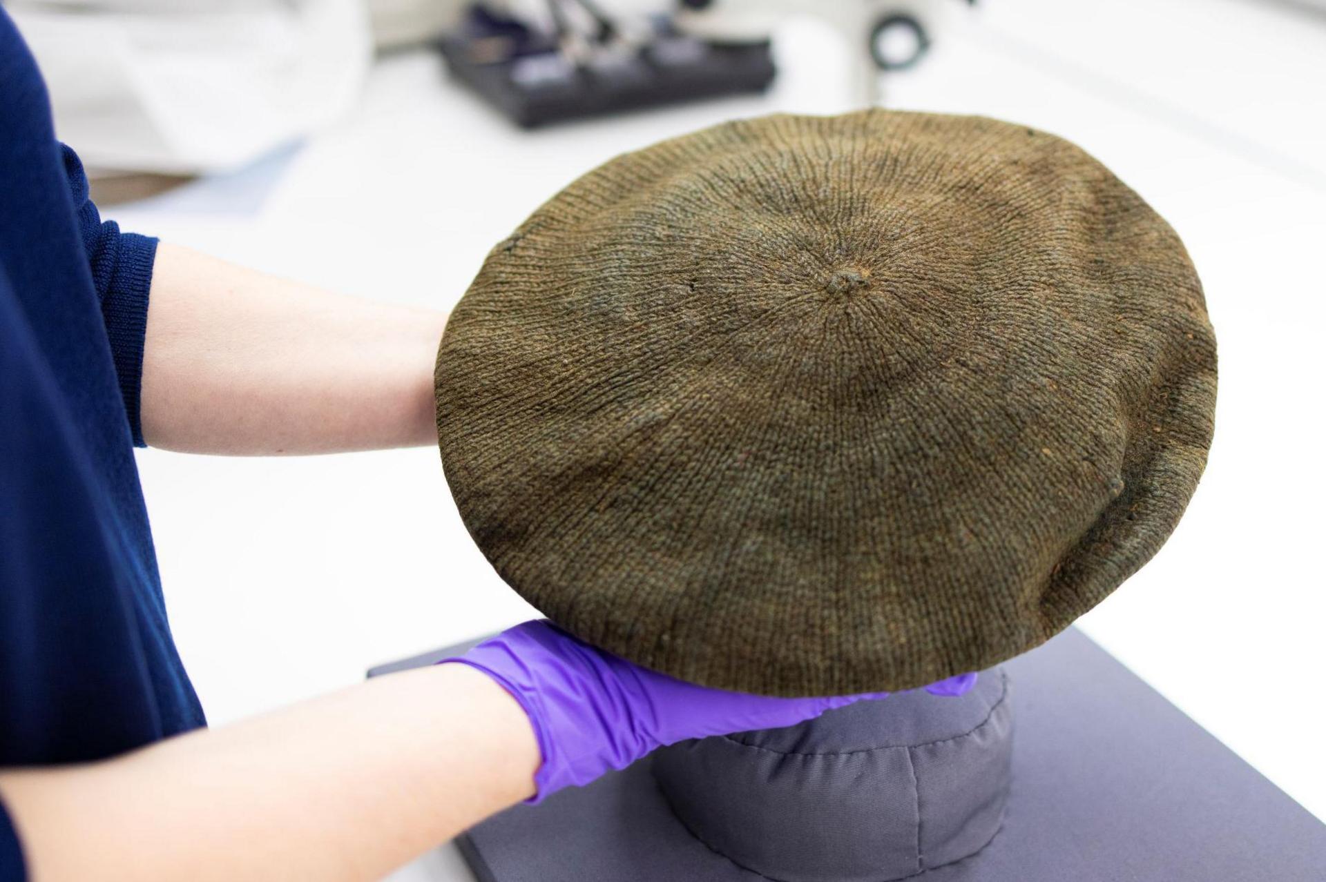 A conservator wearing purple rubber gloves holds up the bonnet. It is a green-brown colour, but was once blue.