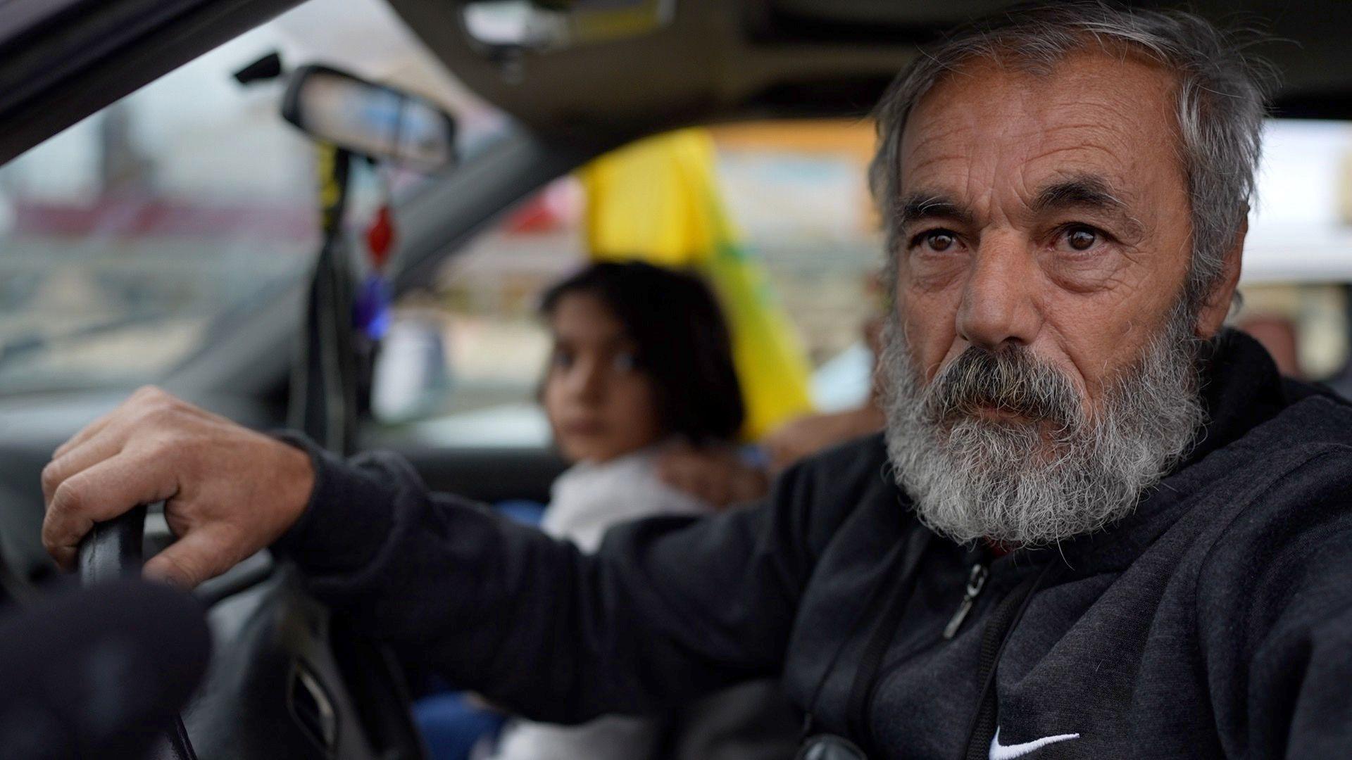 Abu Ali sits in the front seat of his car, hand on the steering wheel. A child and presumed relative sits in the passenger seat next to him.