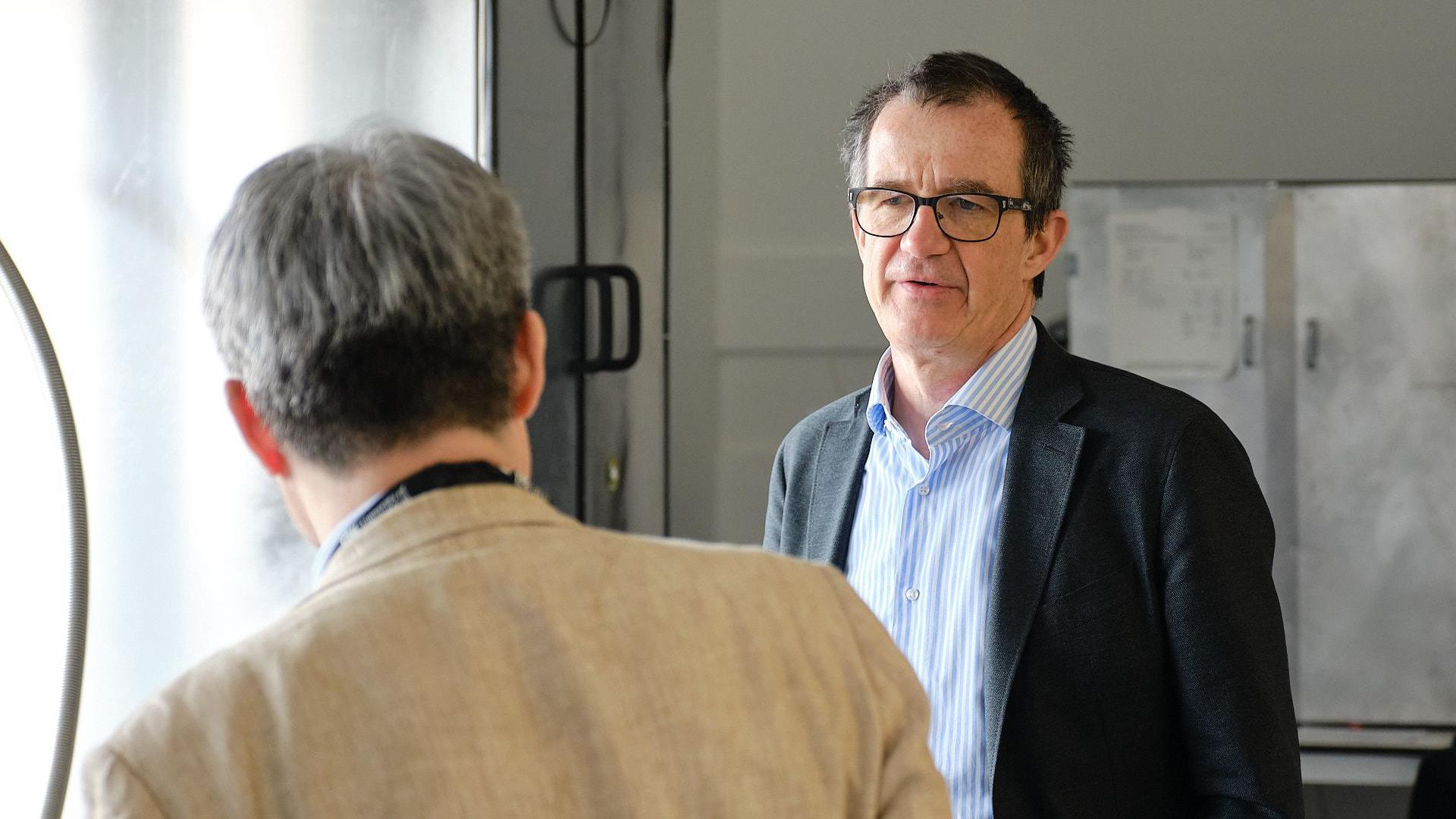 Image shows Professor Martin Kuball talking to a man with his back turned. Prod Kuball is facing the camera, wearing a dark suit jacket and a light blue and white striped shirt. He is wearing glasses.