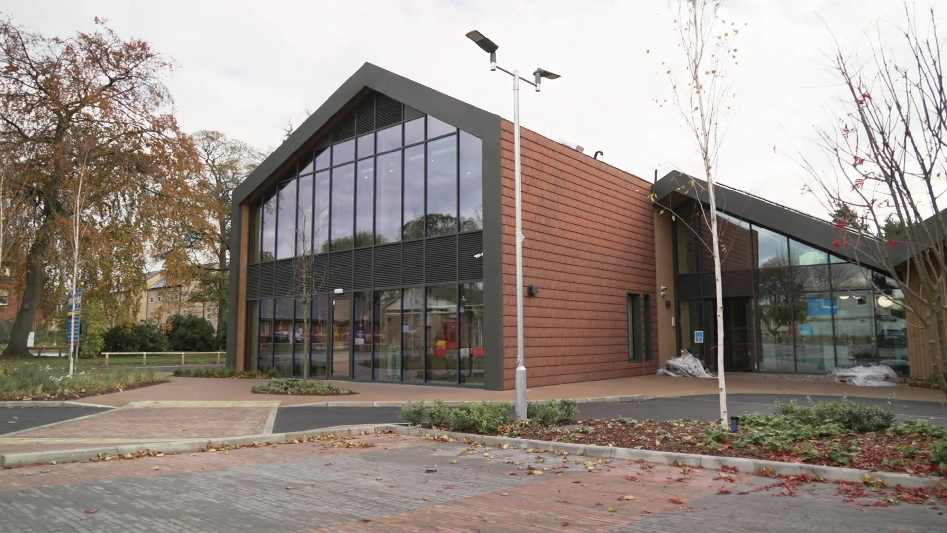 A brown and black building with a pitched roof and glass front. Autumn leaves are in front of the building.