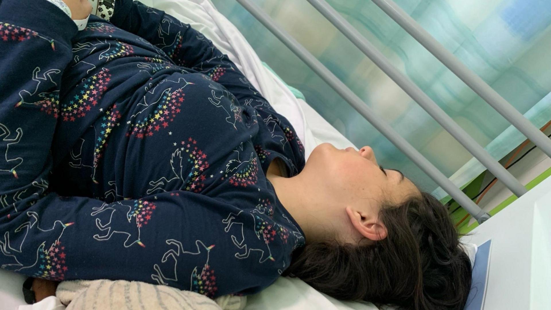 A teenage girl wearing dark navy pyjamas with unicorns on, is seen laying in a hospital bed with her eyes closed.