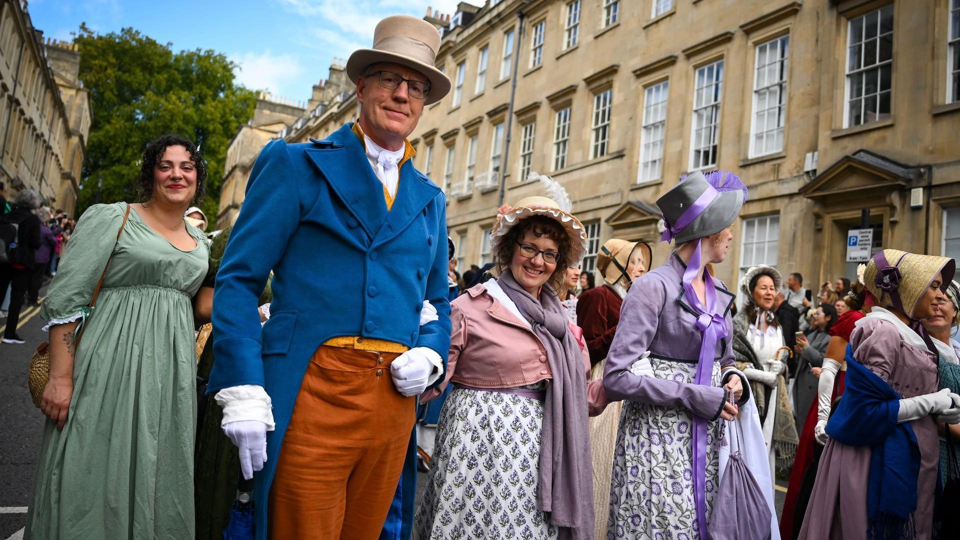 A crowd of people wearing regency costumes walking through Bath. A man wearing a blue suit jacket, brown trousers, a top hat and white gloves is looking at the camera. Two women stood by him are also looking at the camera and smiling. 
