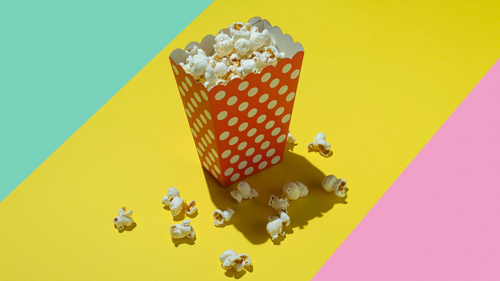A polka dot (white dots on red background) carton of popcorn on bright background of three coloured diagonal stripes which are turquoise, yellow and pink. 