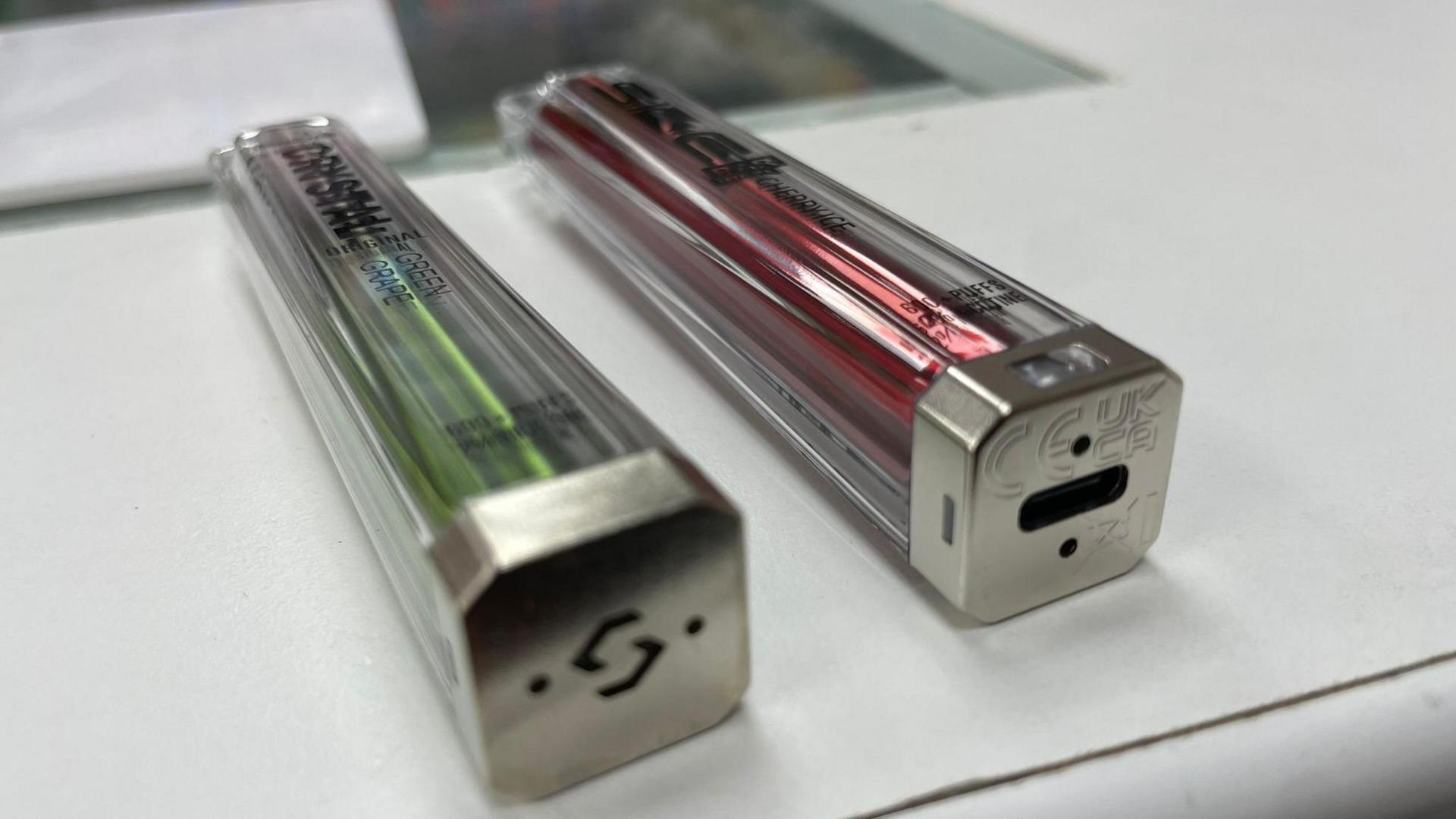 Two vapes next to each other, one is disposable and the other is a rechargeable vape with a charging point at the bottom
