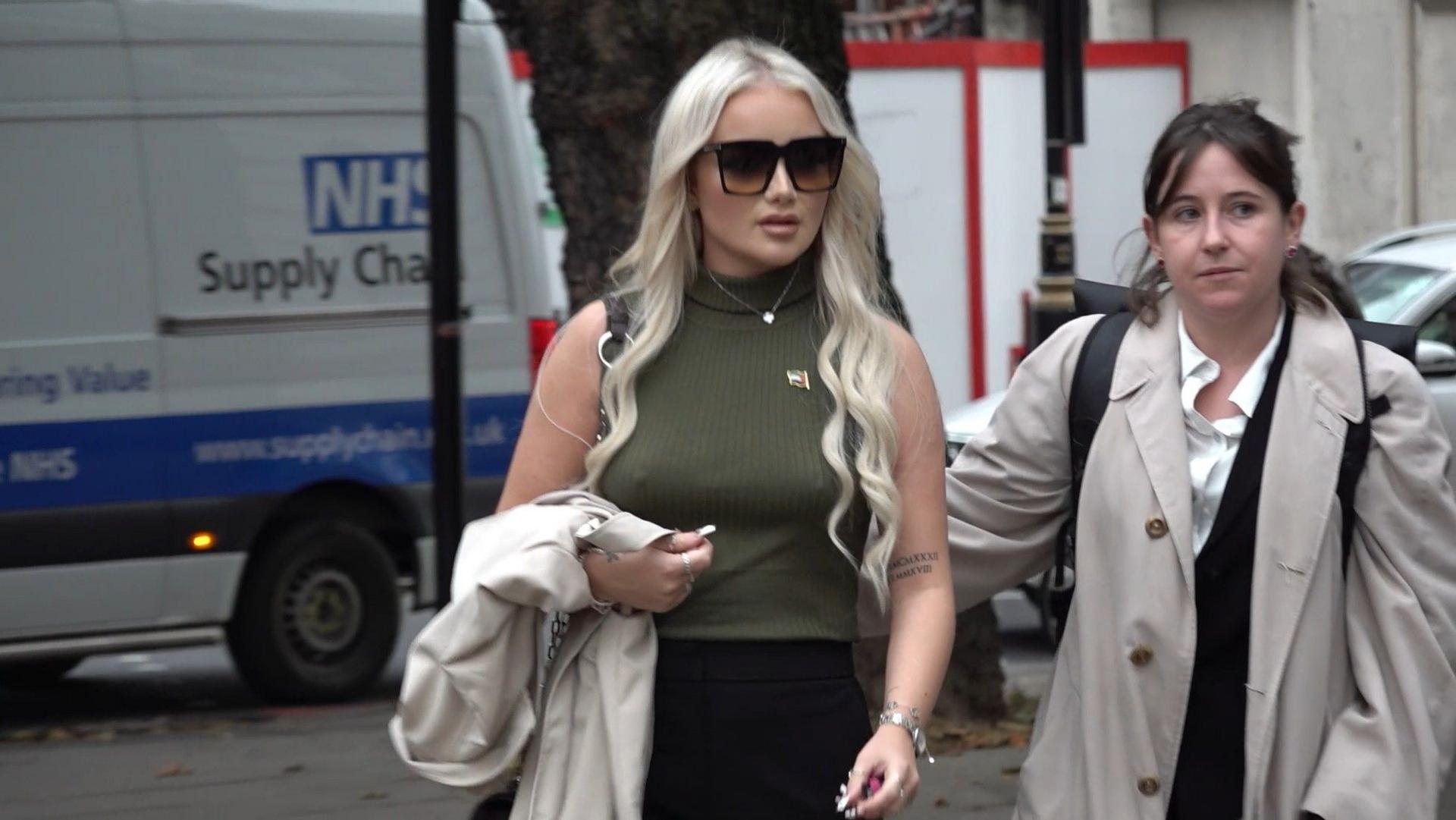 Victoria Thomas Bowen arriving at Westminster Magistrates' Court in London. She has long blonde hair, and is large black wearing sunglasses, a green sleeveless polo neck top and black skirt. She is carrying a light coloured coat. 