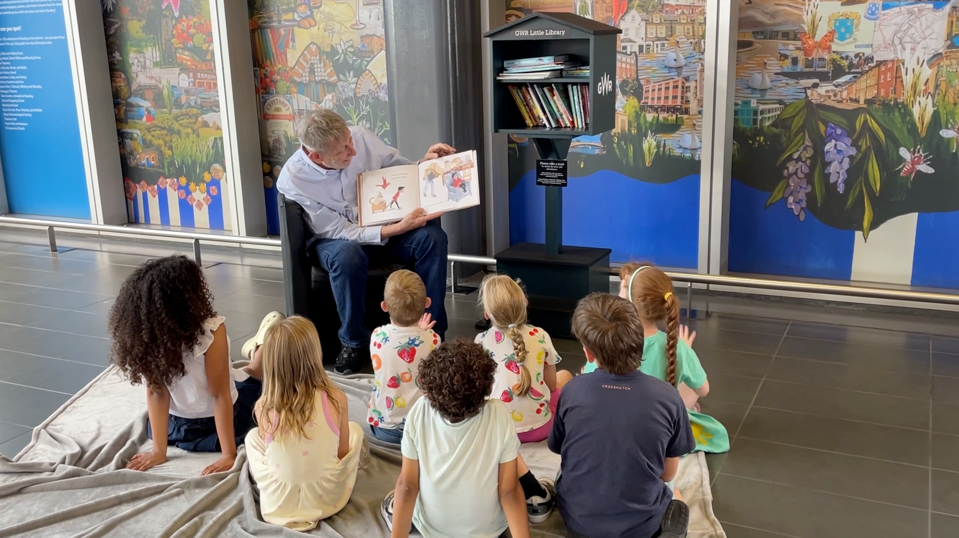 Author Michael Rosen seen reading to a group of children who are sitting next to the GWR Little Library