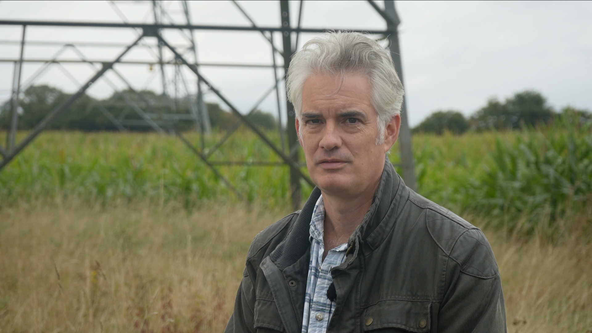 A picture of James Cartlidge. He is in a green field with the base of an electricity pylon pictured behind him. He has grey hair and is wearing a dark green jacket with a blue chequered shirt underneath.