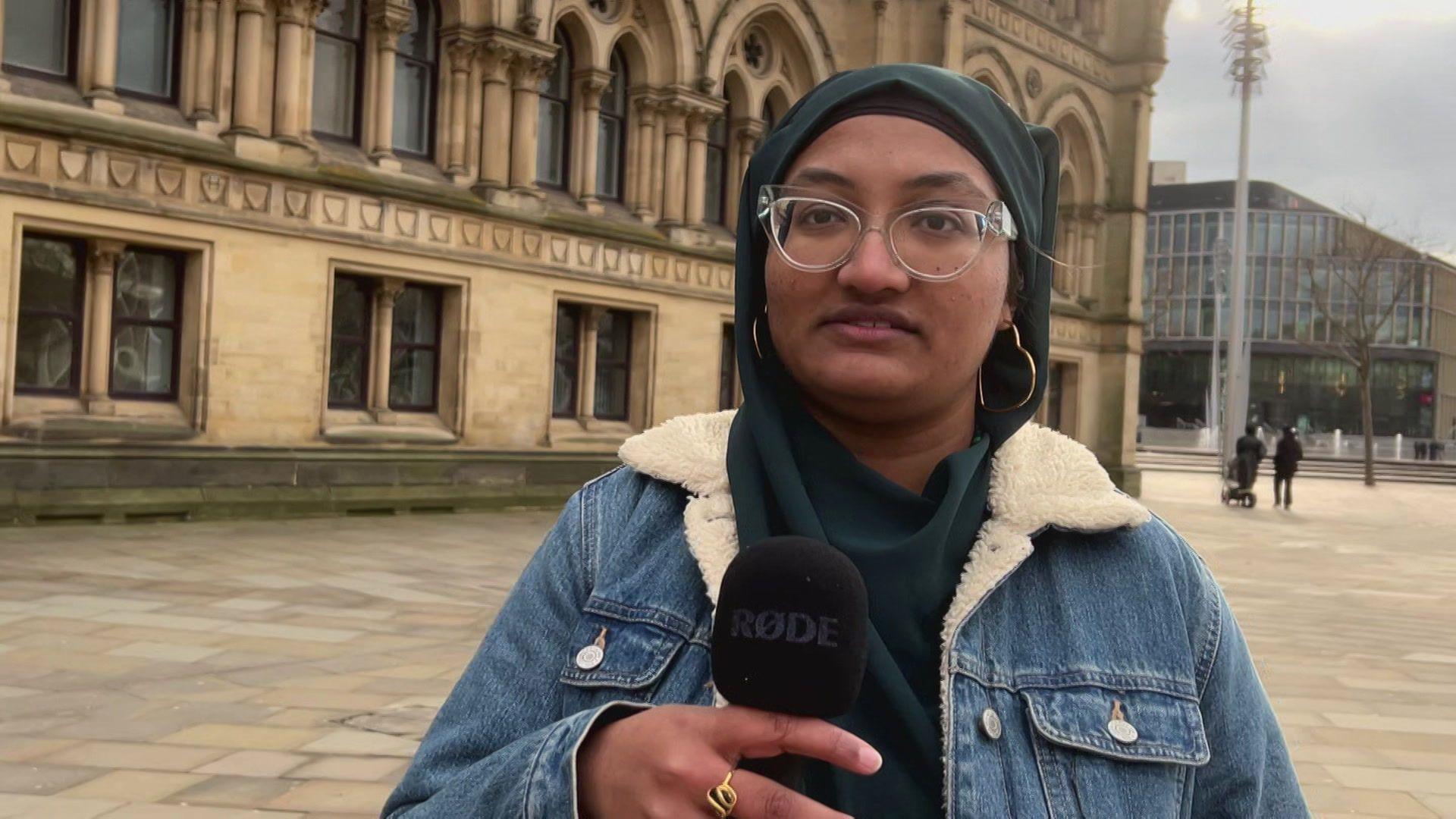 A young woman wearing a blue headscarf and denim jacket. She is standing in front of a historic building in a city square. She wears glasses. She is holding a microphone.