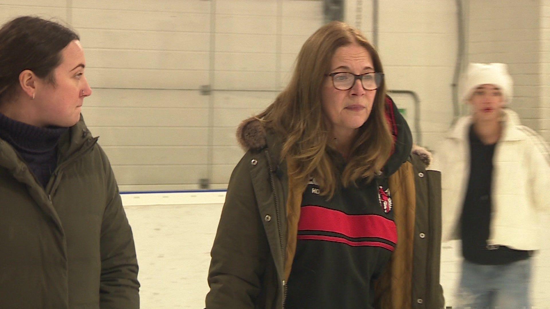 Hannah Driver (right) skating on the ice she's wearing a green coat over a black hoody. She has long hair and glasses. Another skater wearing a white jacket and hat can be seen in the background.