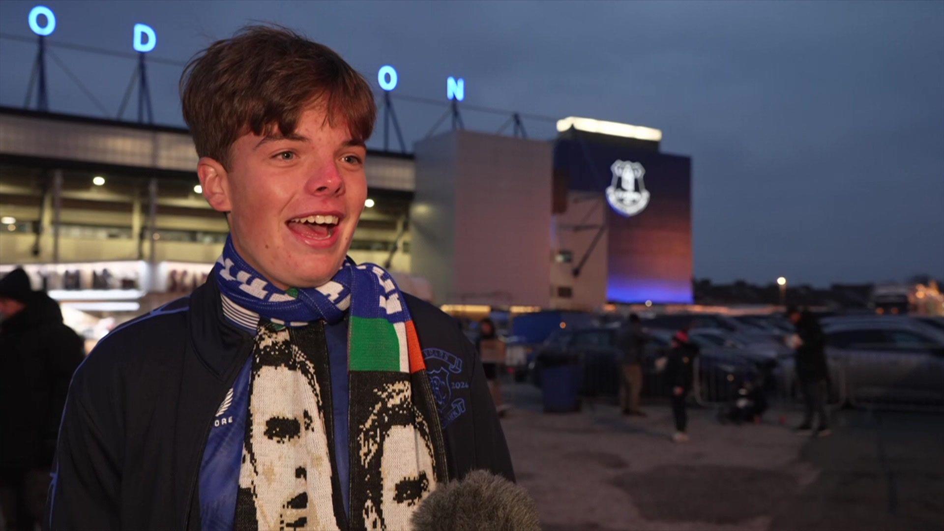 Mackenzie stands in front of Everton's Goodison Park stadium, he has short dark hair and wears an Everton scarf