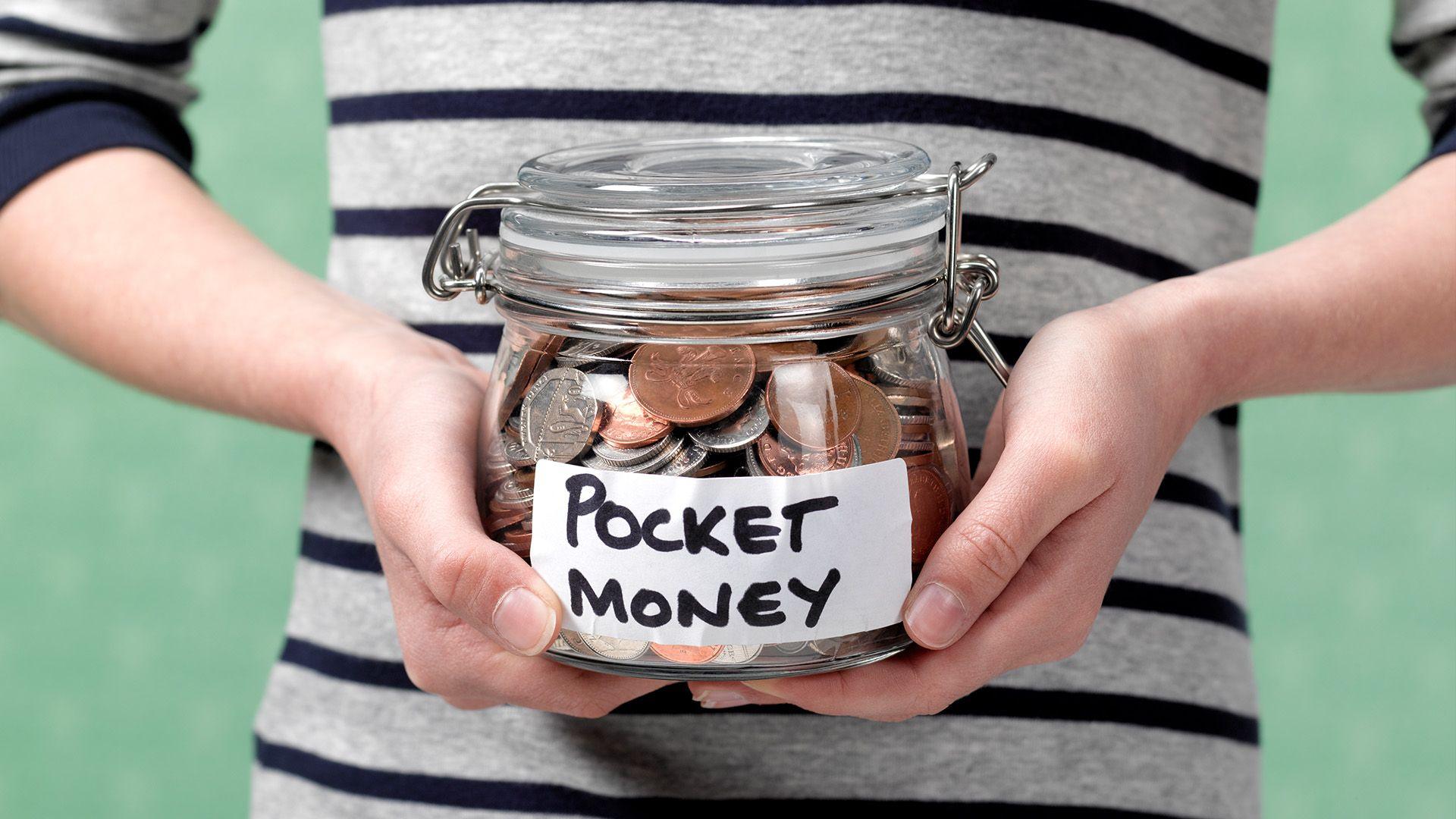 A child is holding a jar with coins in it. There is a label on the jar that says pocket money