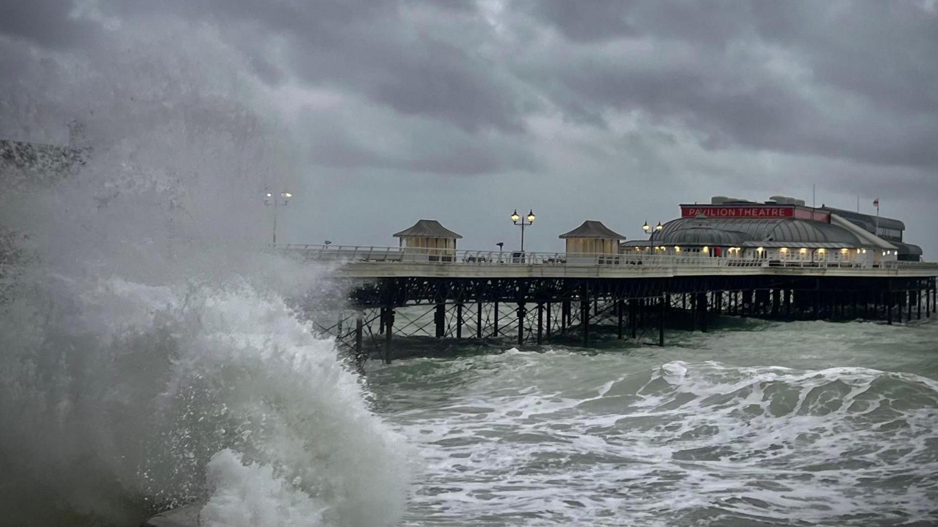 The pier at Cromer with a choppy murky sea below and a huge wave crashing against the wall, at the front of the photo. At the end of the pier is Pavilion Theatre.