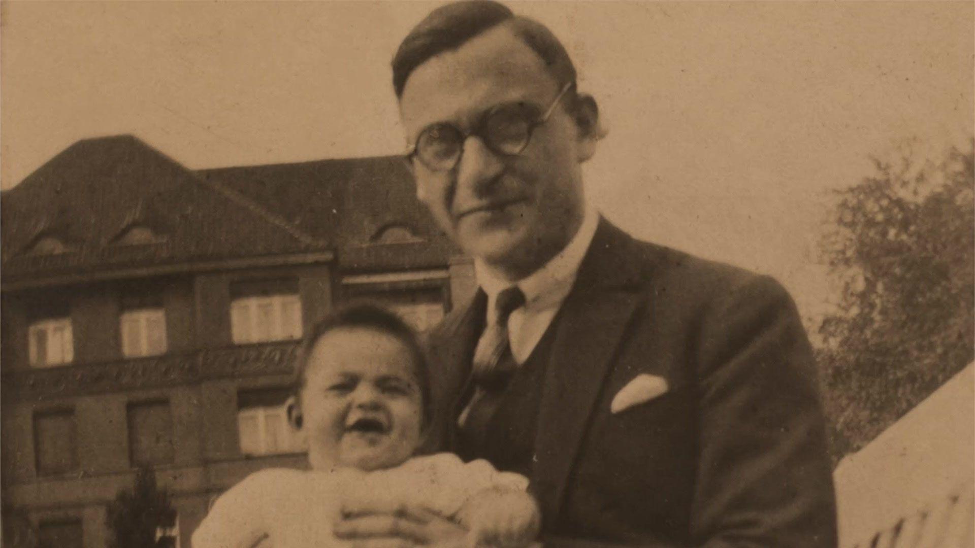 A black-and-white photograph of a bespectacled man in a suit, holding a laughing baby.