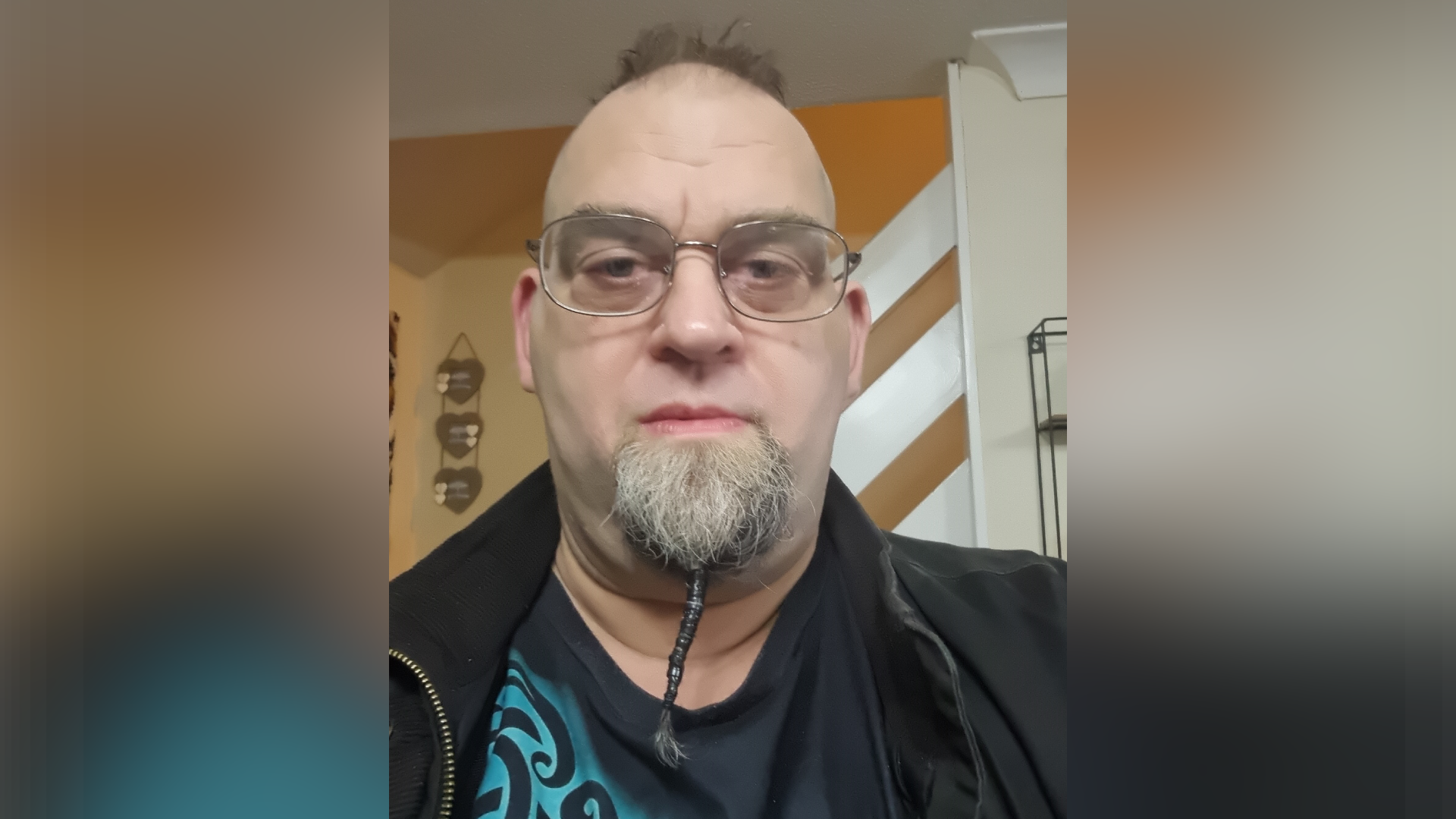 A selfie-style photo of Eddy Shuttleworth. He slightly smiles at the camera. He is largely bald with a small patch of dark hair on his head. He has a grey beard that has been twisted and hangs from his chin. He wears a black jacket with a black and blue t-shirt underneath.