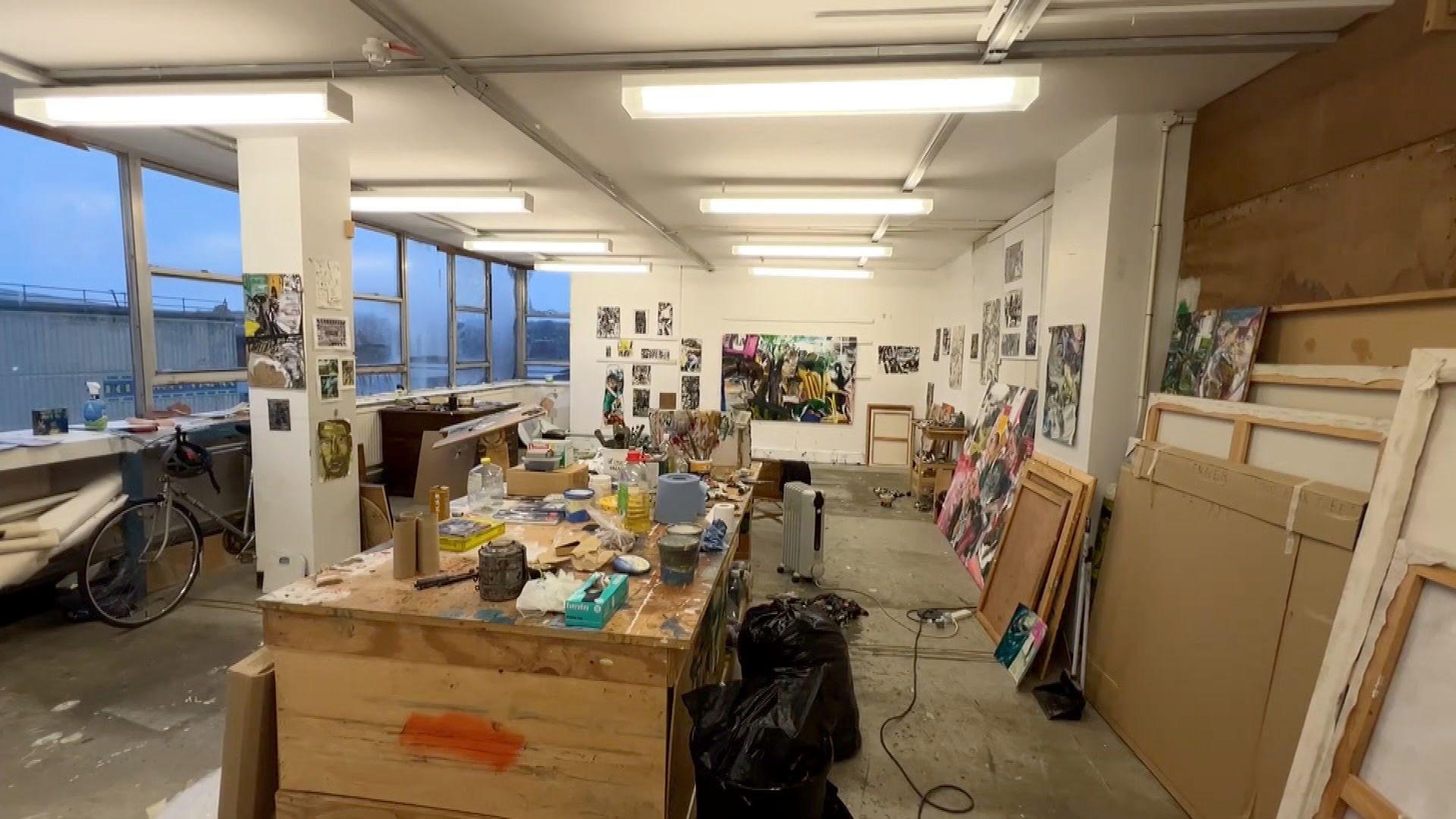 Interior of an artist's studio. There are paintings on the walls and a large workbench with artist materials on. A bike is in the space and lots of canvases have been places along the bottom of a wall. Strip lighting adorns the ceiling. The large room has a concrete floor and an electric heater is near the work bench. 