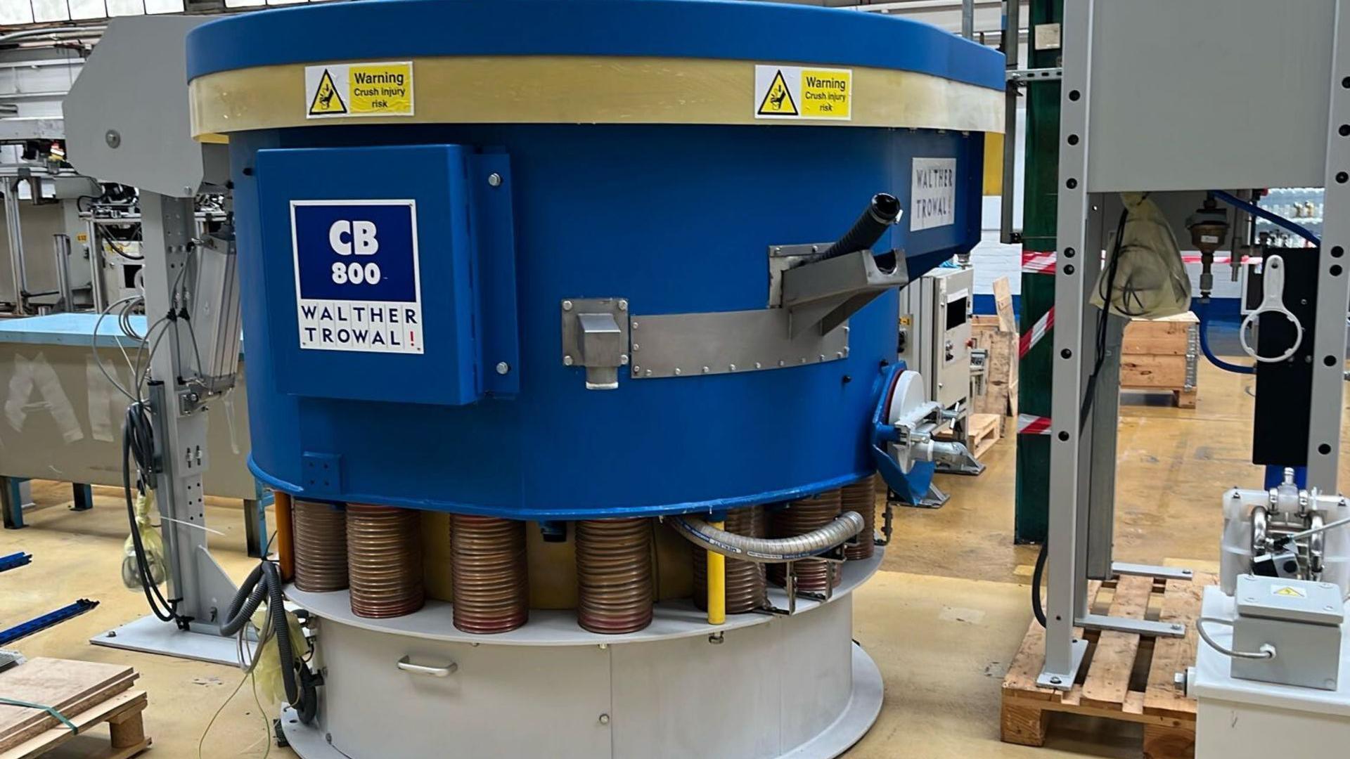 A piece of industrial machinery inside the former SKF factory in Luton. It is cyclindrical and mostly royal blue, with yellow safety warning labels on. The middle section consists of coin-like bronze coloured stacks and the base is white. It is labelled CB800 Walther Trowal. There are other machines and hazard tape in the the background and the floor is a light wood.