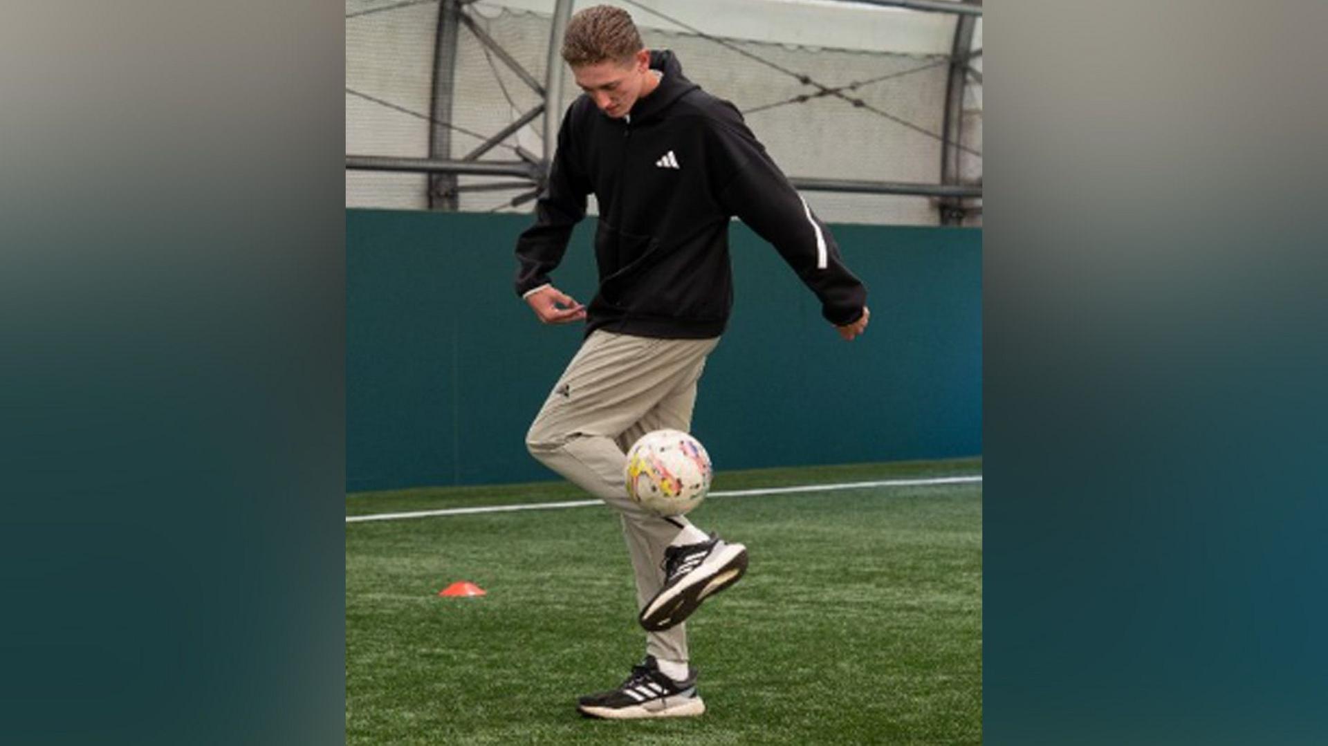 A young man with blond hair wearing a black jacket and light trousers kicks up a football. He stands on astroturf.