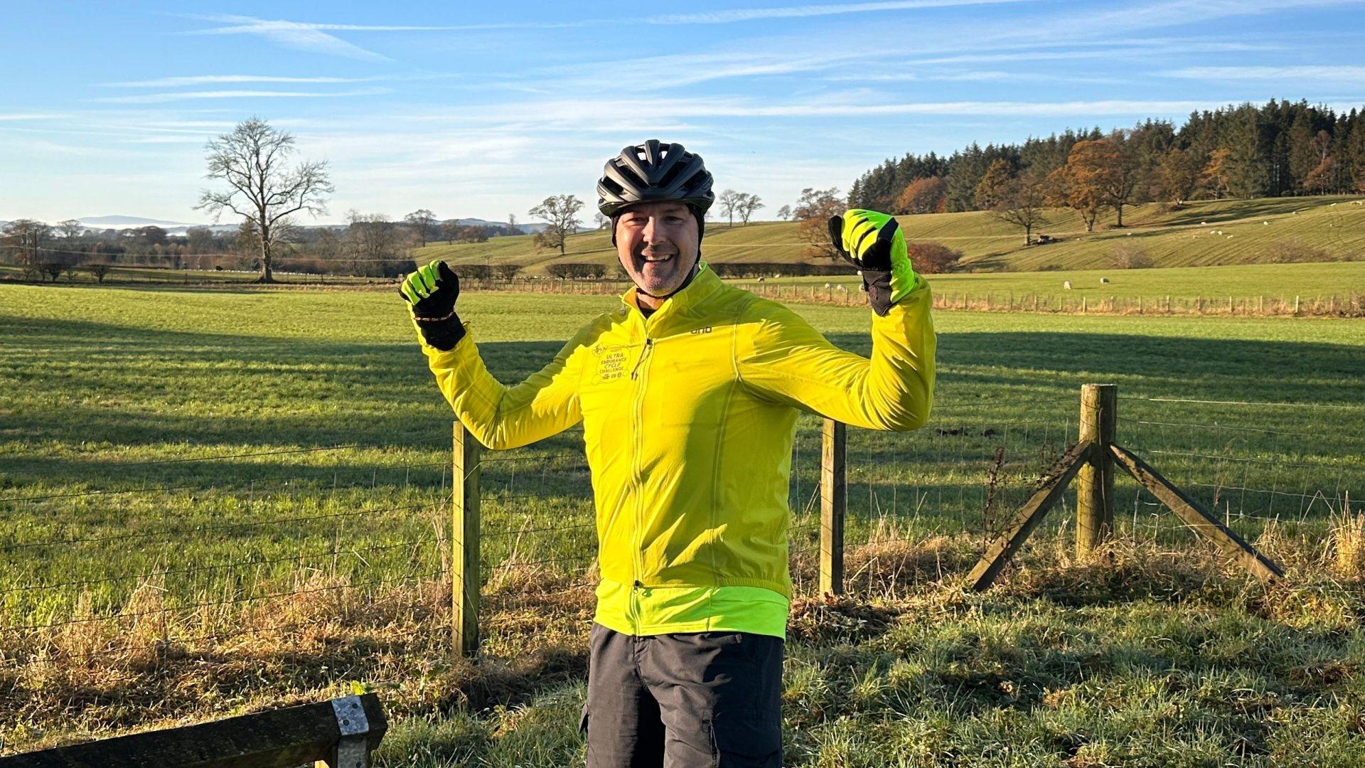 Paddy McGuinness raises his arms triumphantly wearing yellow cycle gear and a big smile on his face, in front of blue skies and green fields