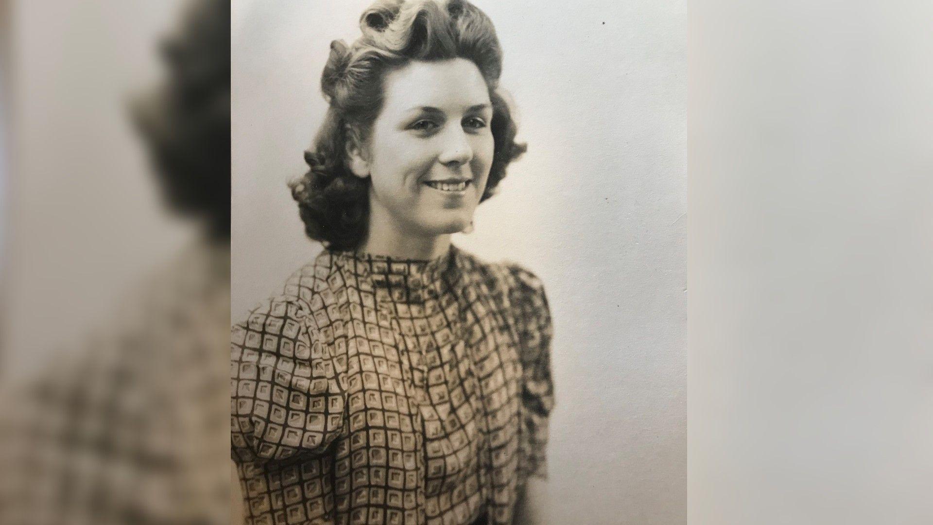 A black and white photograph of Mrs Bond when she was younger. She is wearing a high neck blouse with a square pattern print on it. She is smiling and glancing at the camera with her hair down but styled into position