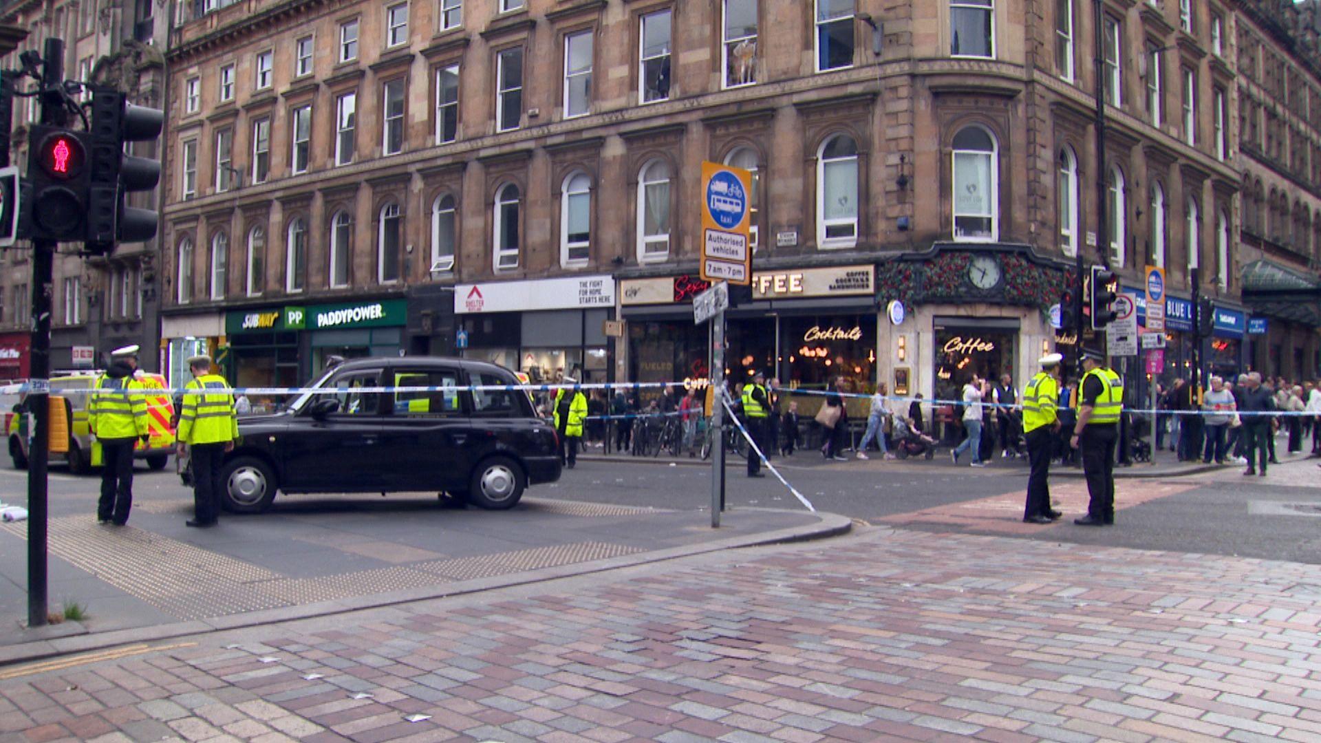 Police officers and shoppers with a black cab visible in the foreground behind police tape