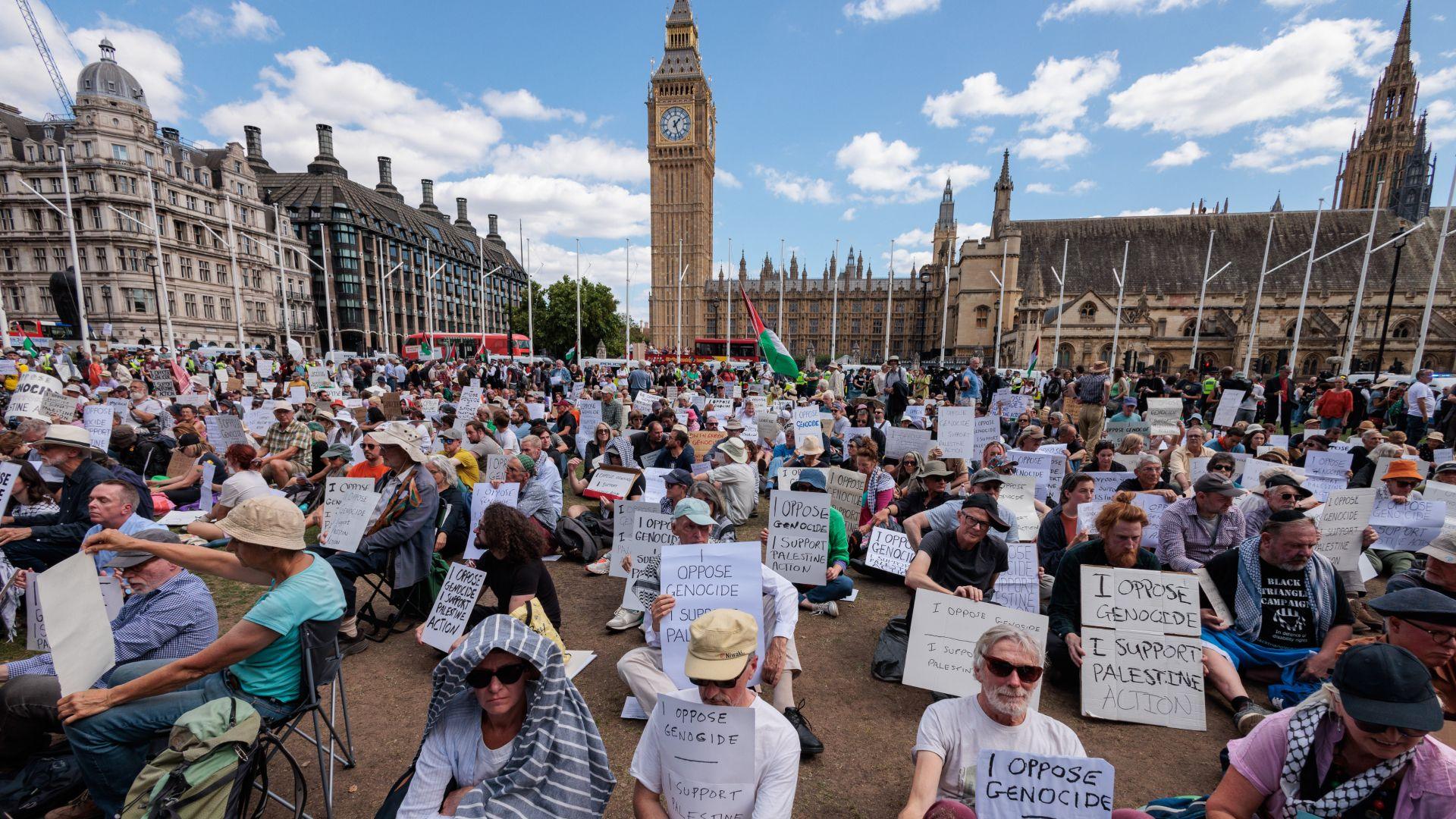 Hundreds of people hold signs reading 'I Oppose Genocide - I Support Palestine Action' while sitting in Parliament Square. Big Ben's clocktower is seen in the background as well as the Palace of Westminster.