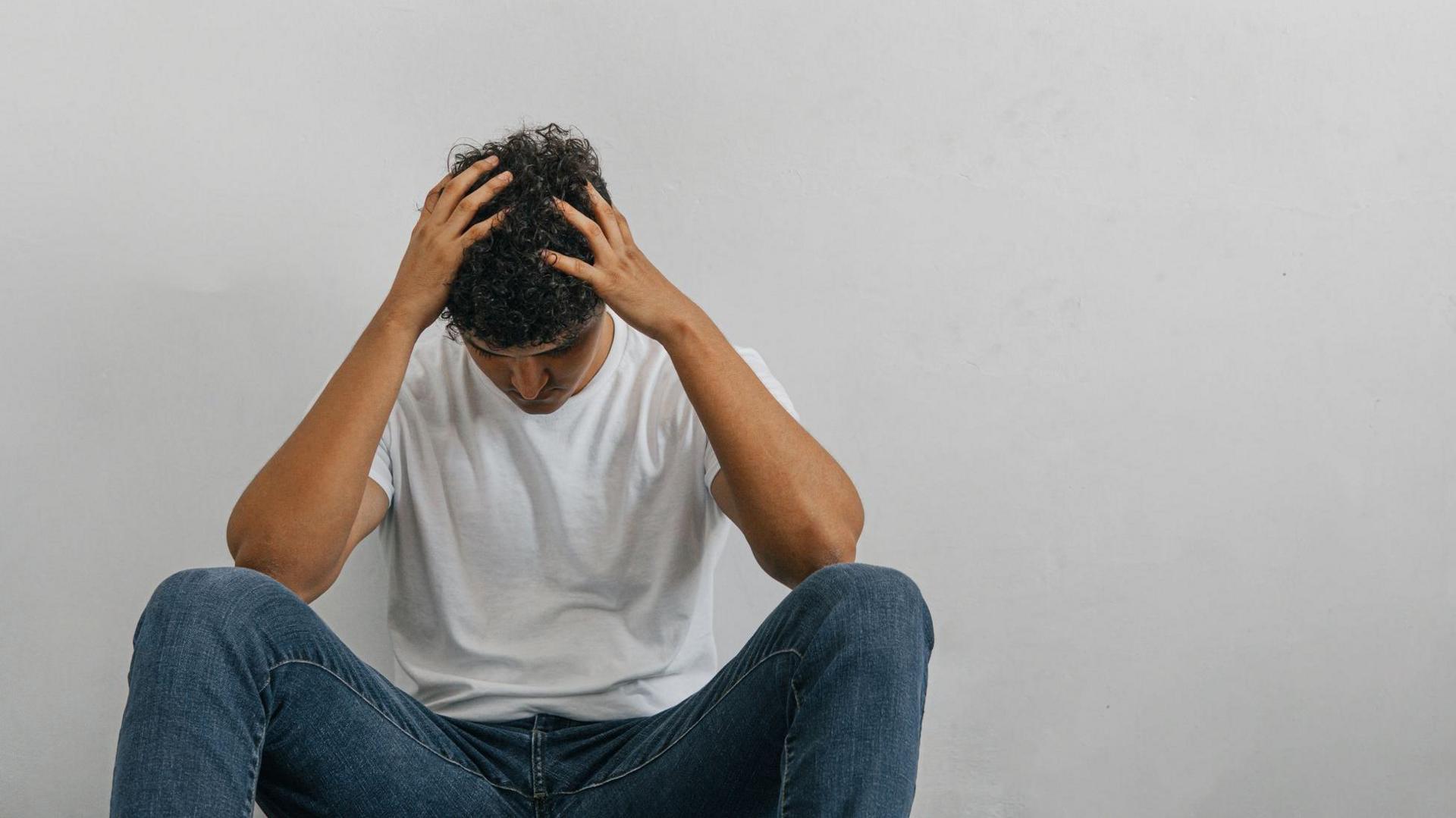 A man in a white T-shirt and jeans sits against a wall with his hands on top of his head looking distressed. 