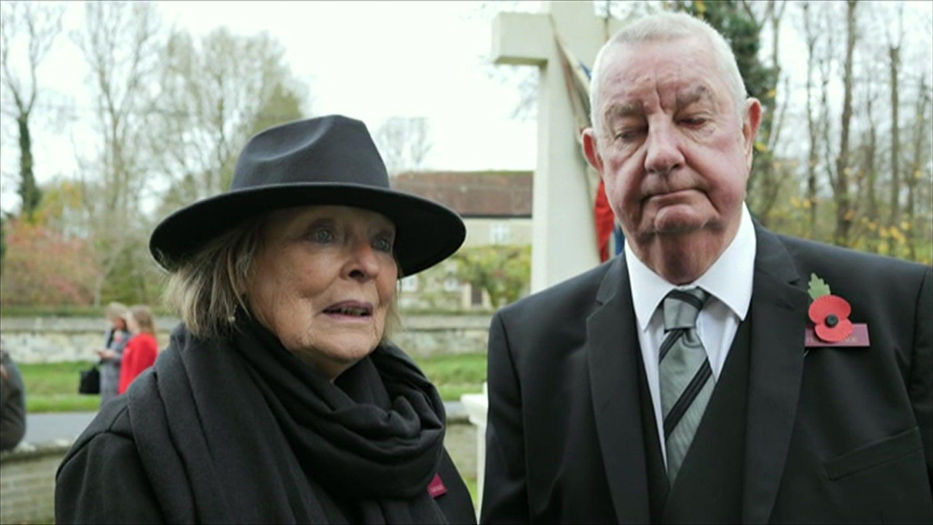 A man and a woman wearing dark formal wear and poppies stand next to each other. The woman is also wearing a black wide-rimmed hat as the pair stand in front of a memorial cross with trees and building in the background.