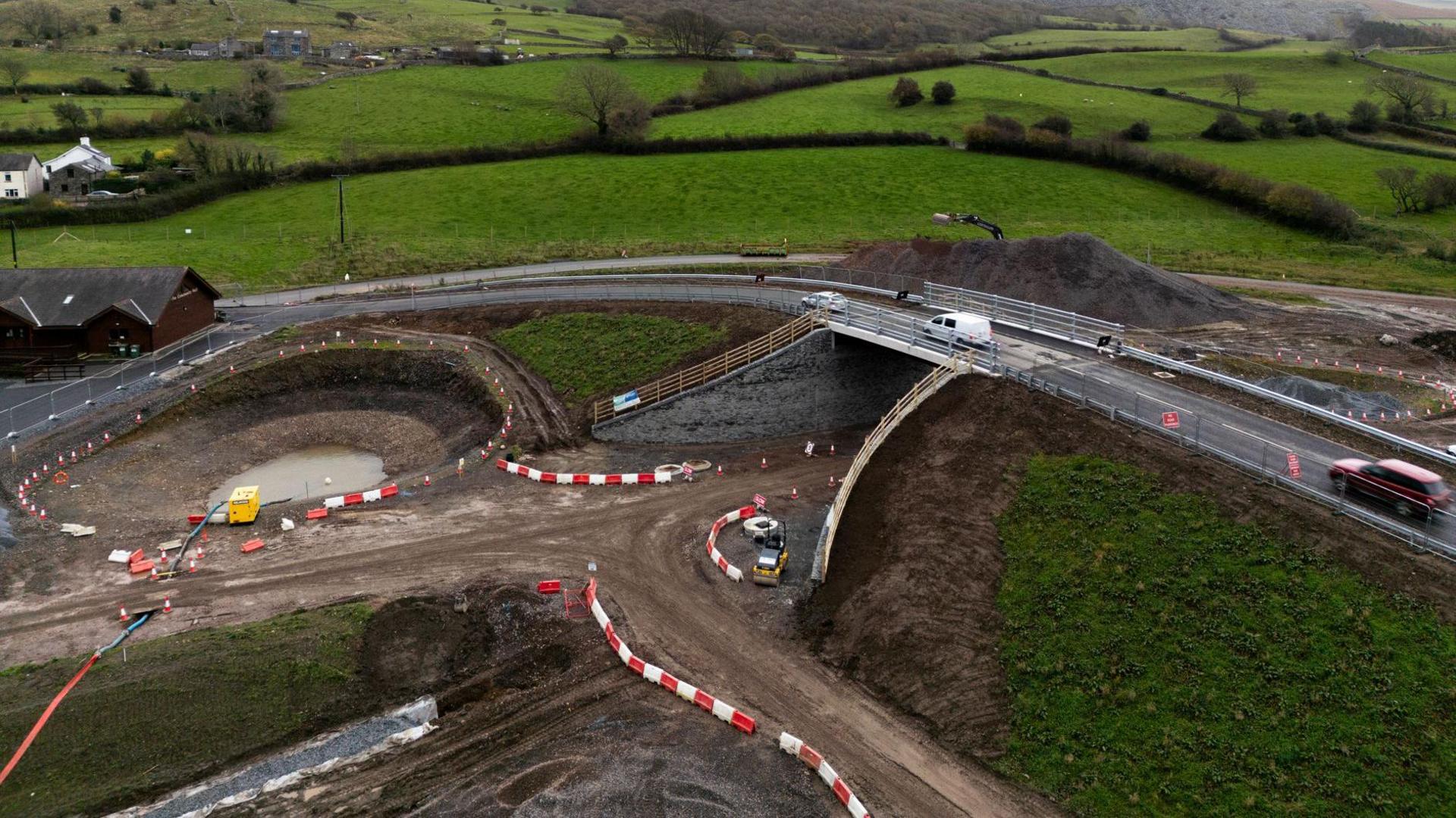The new bridge with cars in motion travelling on it. The bridge is over an unfinished stretch of road, which looks like a dirt track with white and red bollards and machinery on the site. To one side there is a large hole, surrounded by road cones, which has water in the middle of it.