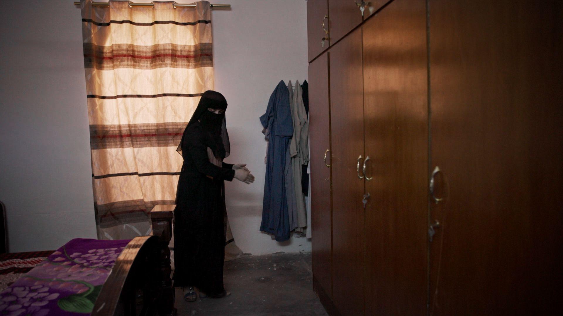 Safa wears a black headscarf, veil and robe covering everything except her hands and eyes. On the left is the foot of a double bed and on the right are some fitted wooden wardrobes. She points at the floor by the far end of the cupboards.