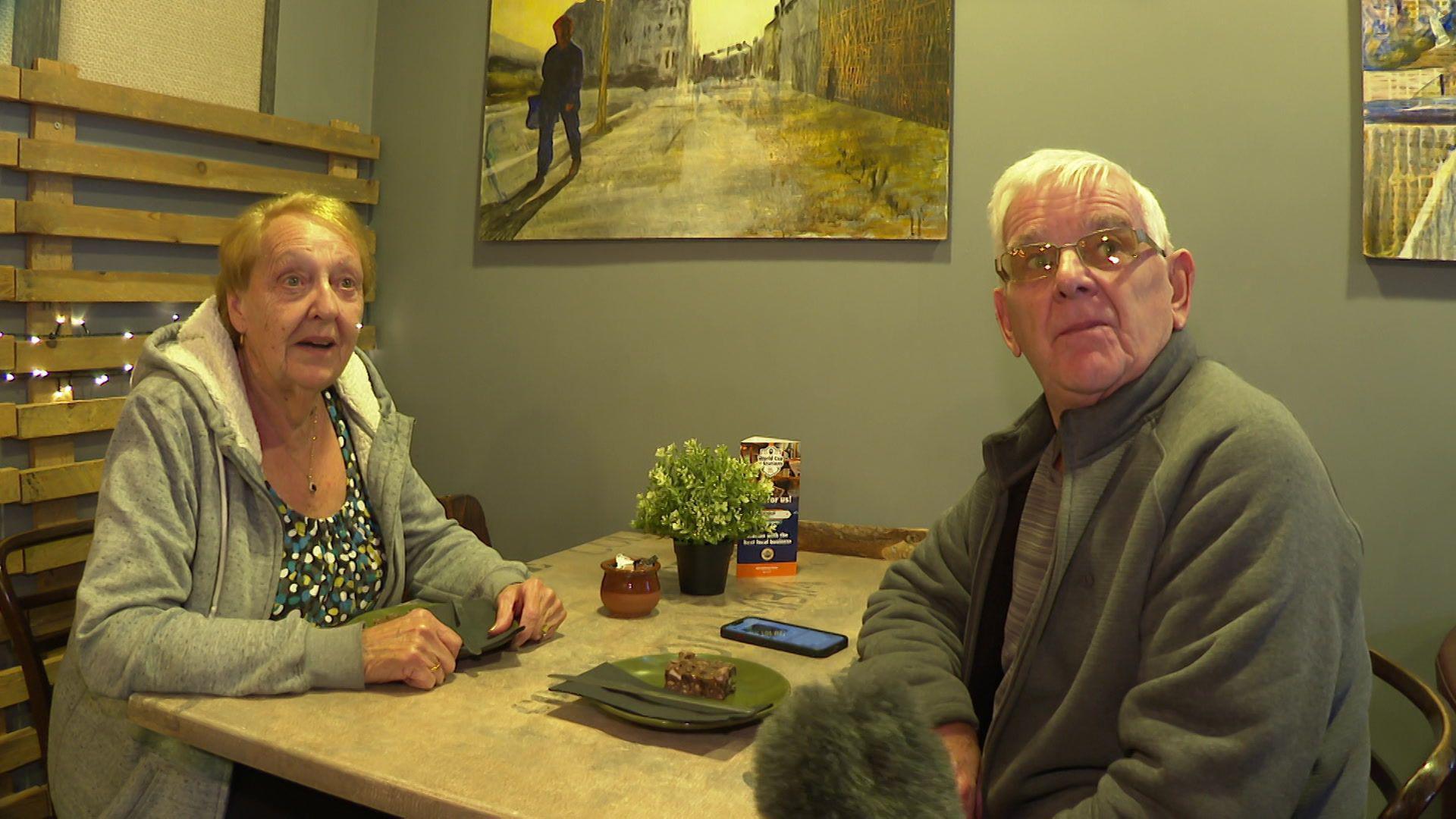 A woman with grey hair, a patterned blue top and a grey zipped jacket sits at a table with a man with grey hair and glasses who is wearing a grey jacket. A slice of chocolate cake is on the table, along with a plant in a plant pot, a sugar bowl and a mobile phone.